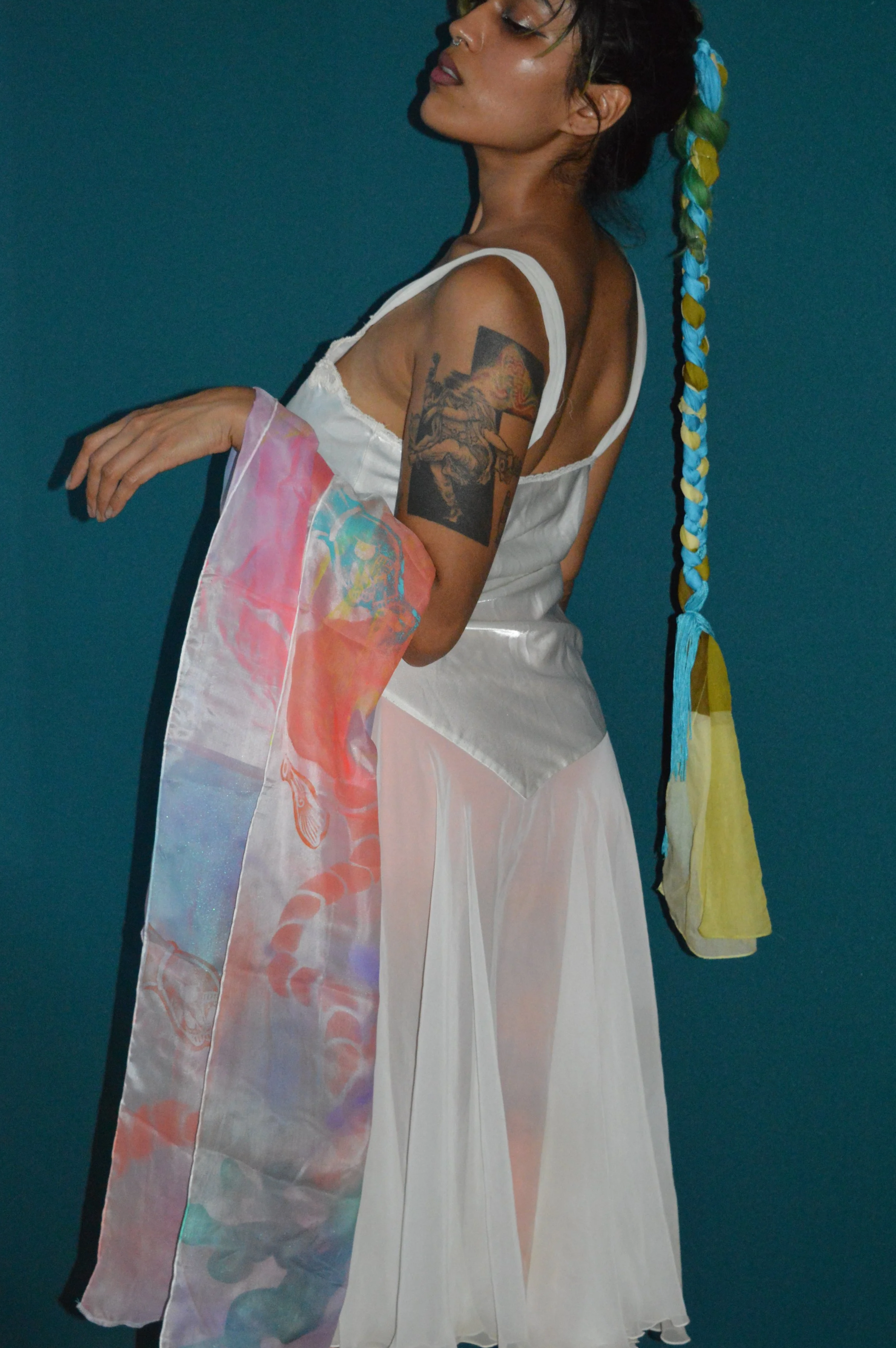 A woman with dark hair styled in a braid with multicolored ribbon, wearing a white dress with thin straps, holding a multicolored scarf in front of a plain blue background.