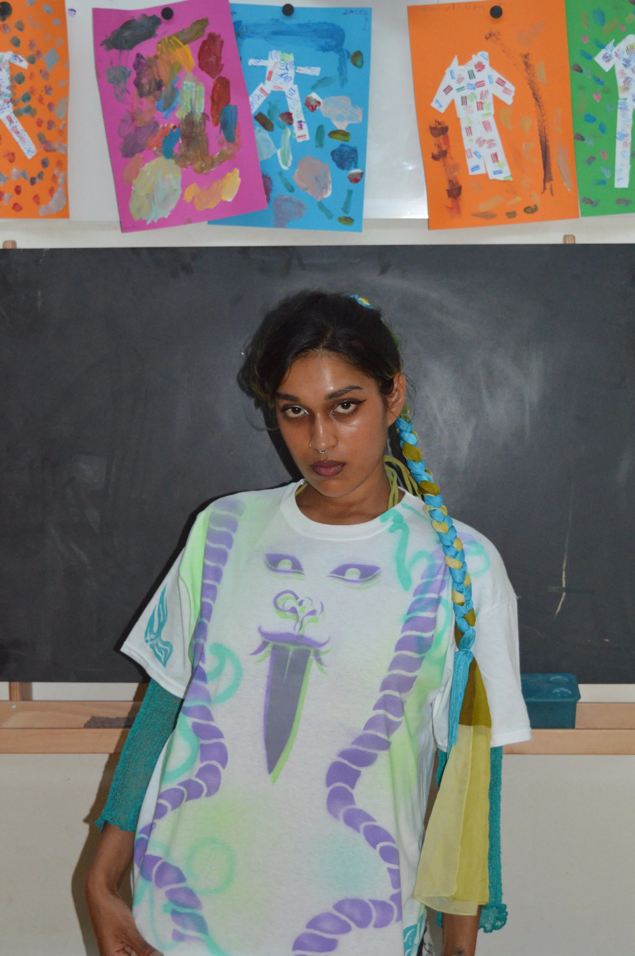 A young woman with dark hair and a braid is standing in front of a blackboard with colorful paintings of abstract figures on colored paper hanging above her. She is wearing a white t-shirt with a design of a snake and a dagger. There is a person in t