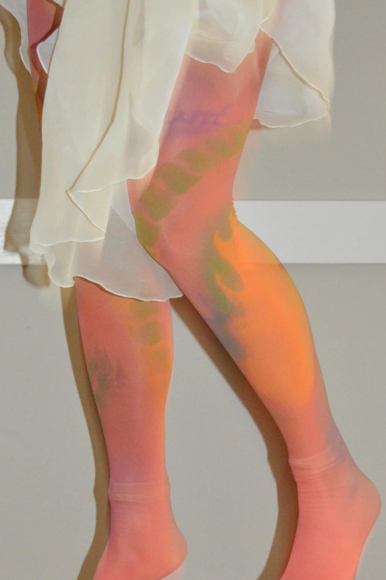 Close-up of a person's legs wearing colorful tights and a white flowing dress, standing against a plain wall.