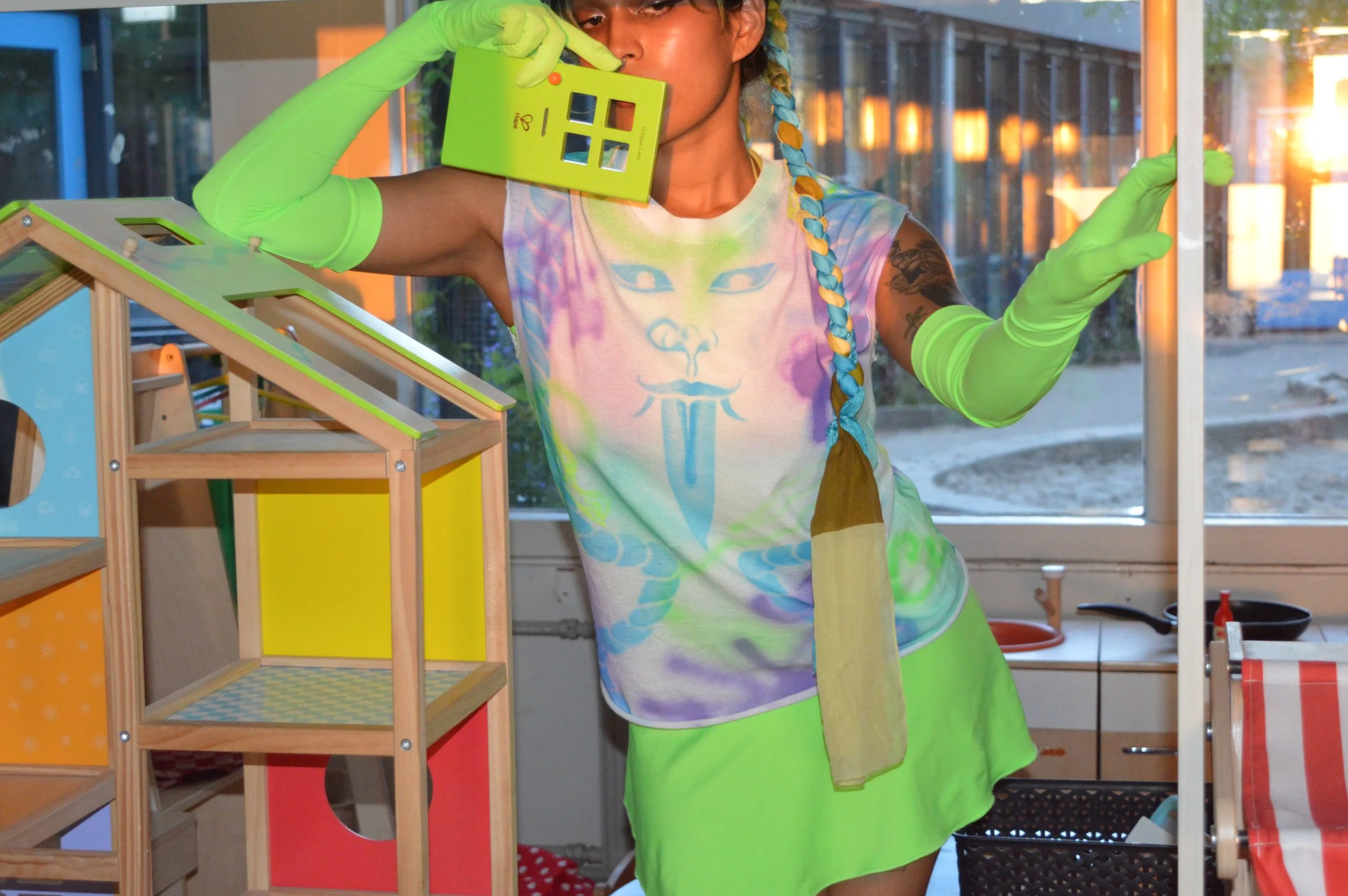 Person dressed in colorful tie-dye shirt, green skirt, and matching green gloves holding a house-shaped object near her face, standing indoors with colorful decor and large windows showing an outdoor view.