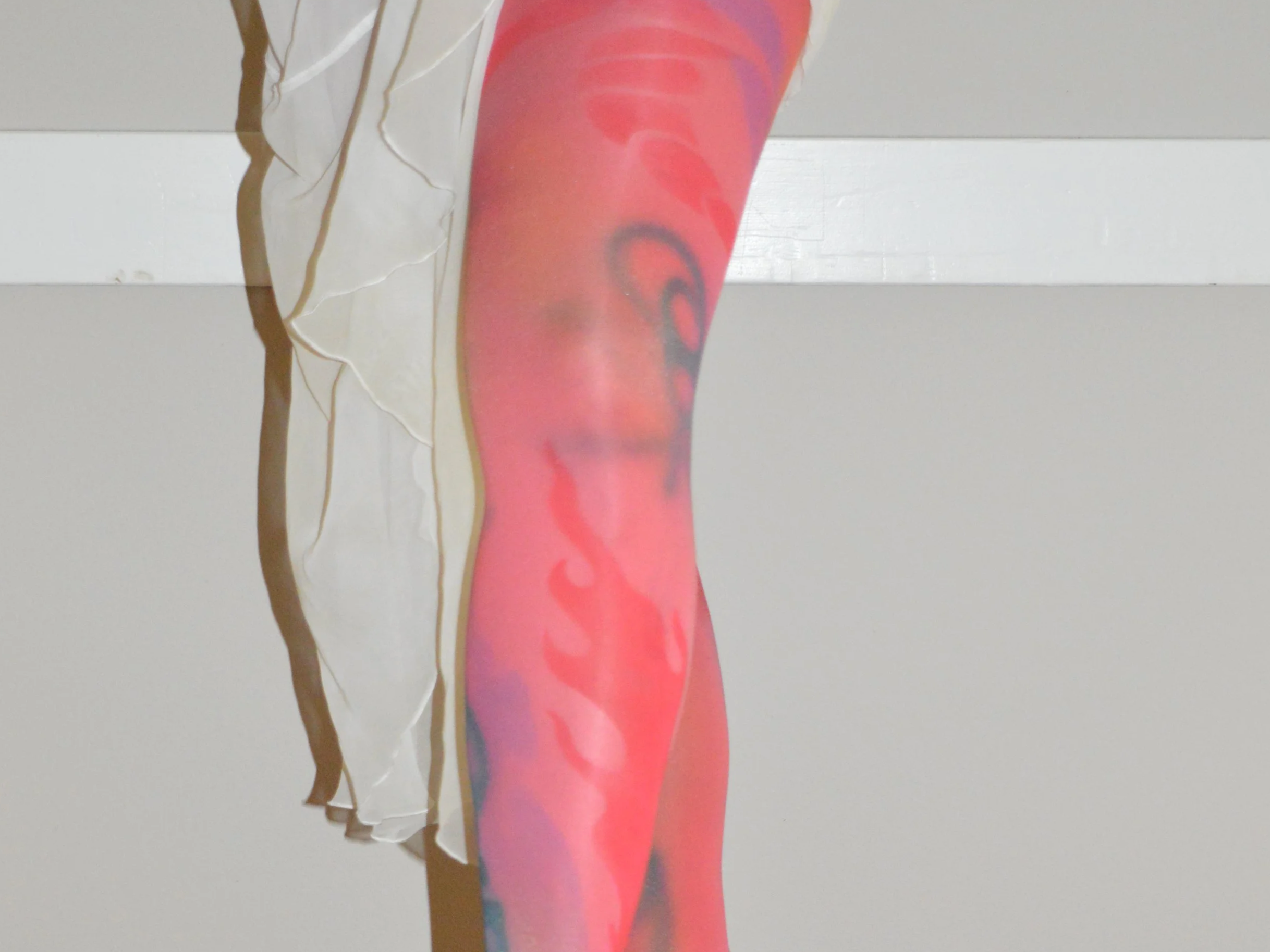 Close-up of a colorful tattooed leg with pink and purple hues, exposed beside a beige, shredded fabric drape, against a plain background.