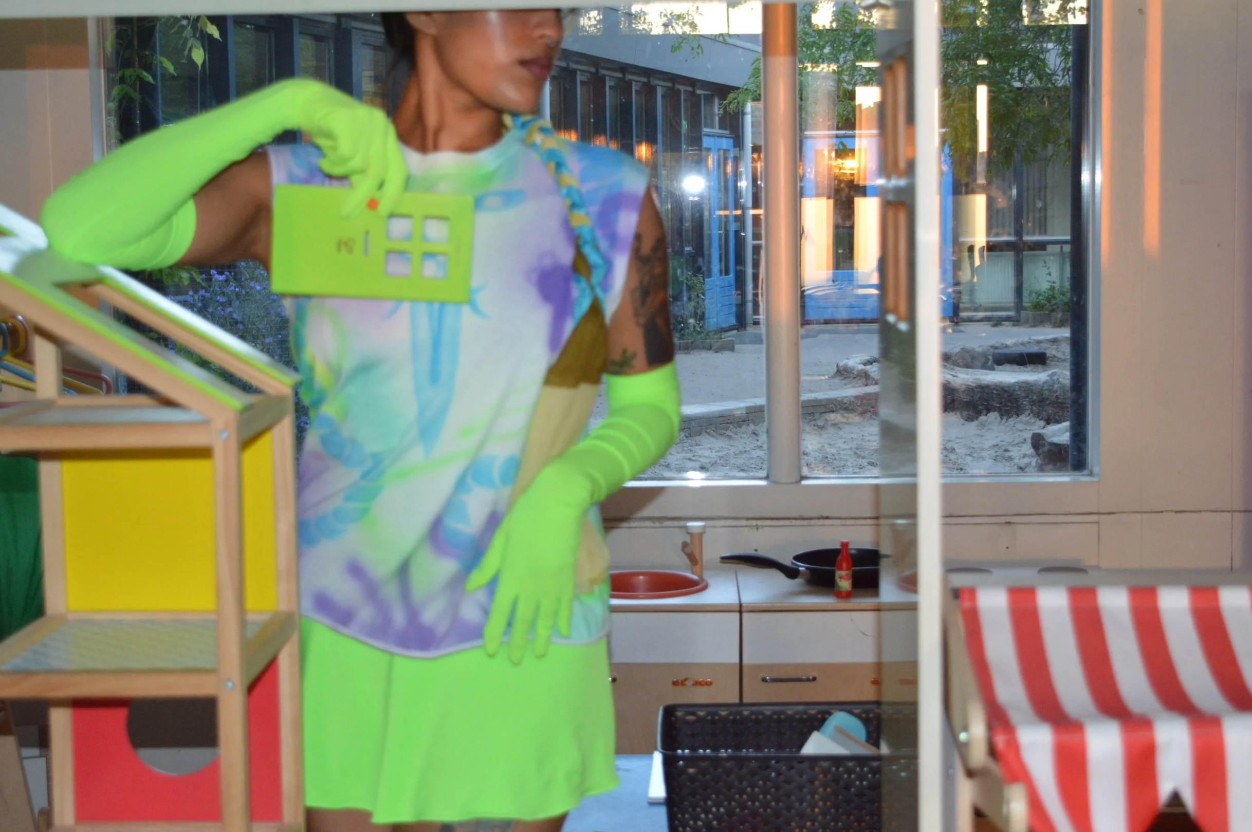 A person dressed in neon green gloves, a colorful tie-dye top, and a yellow skirt is holding a small, green plastic house with a window design on the front. They are standing inside a room with a window showing an outdoor area with trees and a buildi