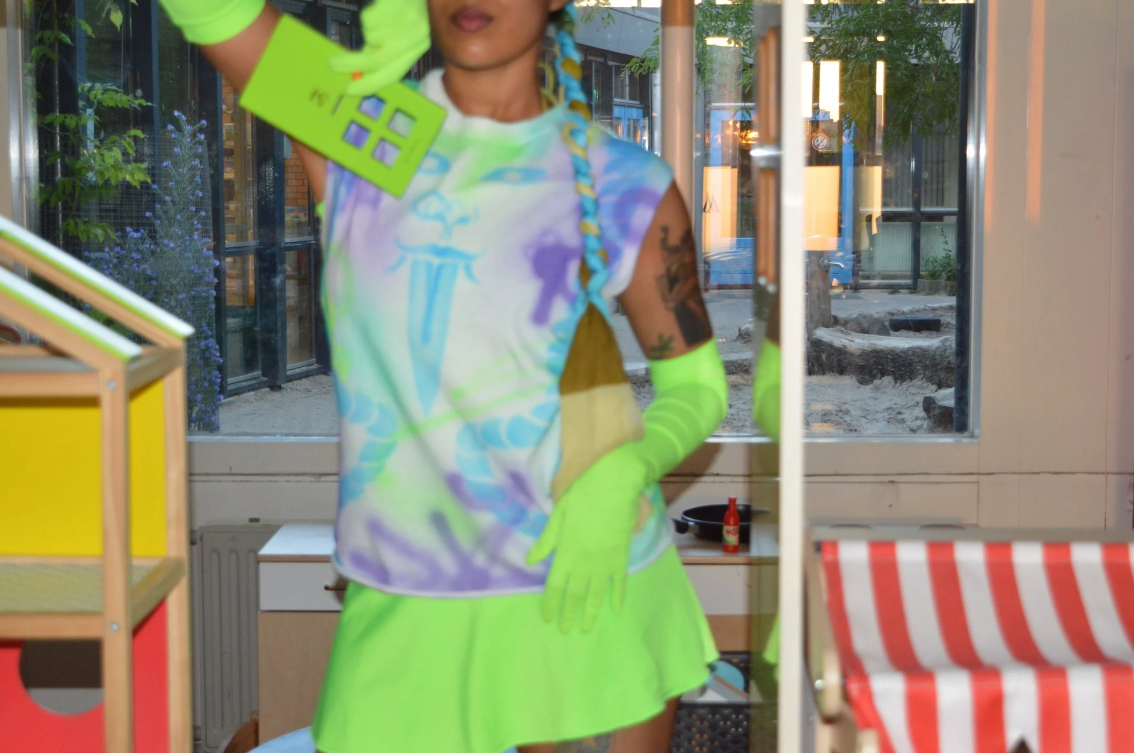 A person behind a glass door, wearing a colorful tie-dye shirt, bright green gloves and skirt, and with blue and yellow braided hair. The person is holding a green card with house icons. Outside the window, there are trees and a building in an urban 
