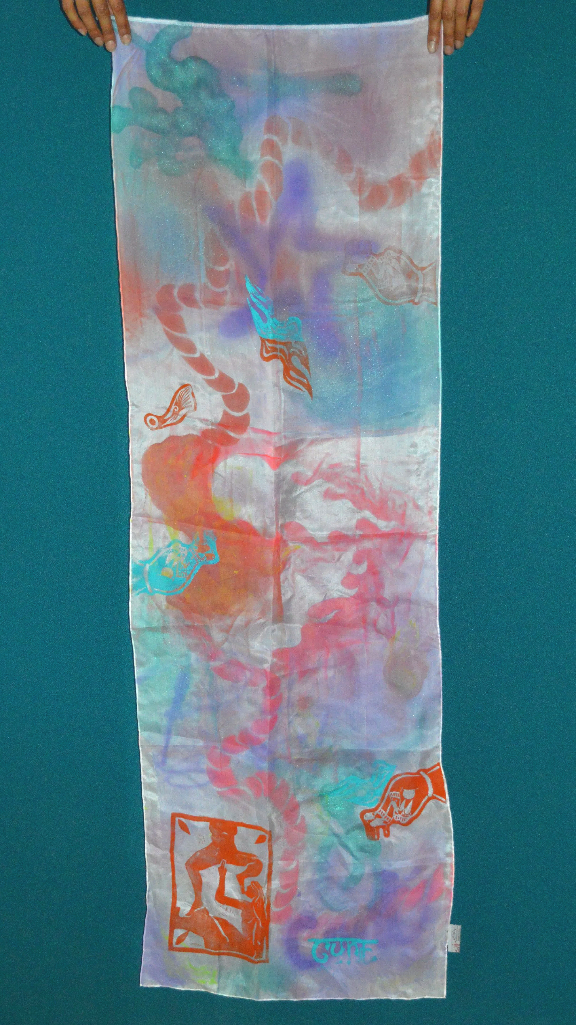 A colorful, semi-transparent silk scarf held up by two hands with a blue background. The scarf features abstract, flowing designs with patterns resembling snakes, skulls, and geometric shapes in shades of orange, blue, purple, and green.