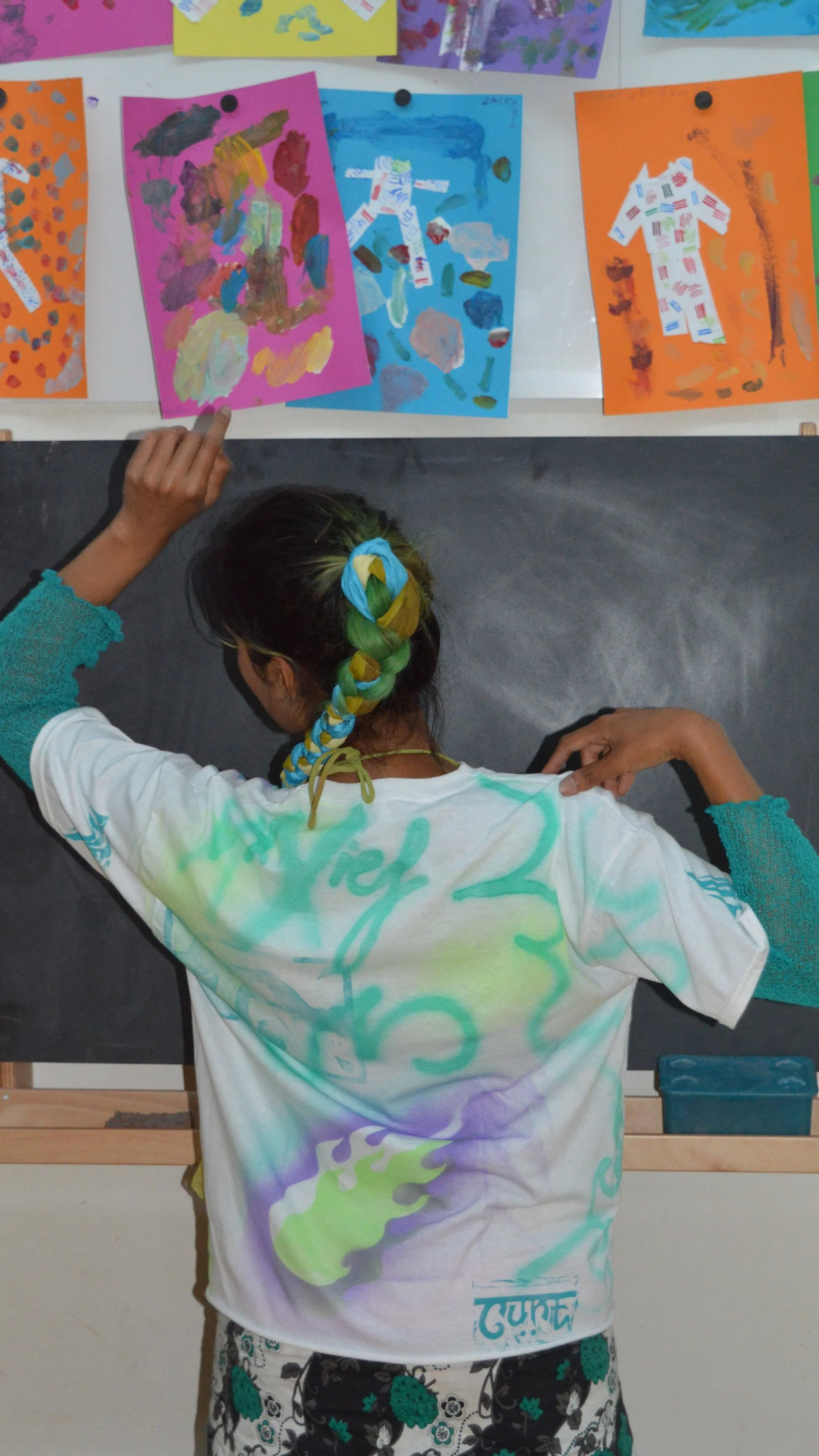 A young girl with a colorful braid in her hair, wearing a white T-shirt with green and purple designs, is pointing at colorful artwork displayed on a bulletin board. The artwork consists of painted and collaged pieces on various colored papers.