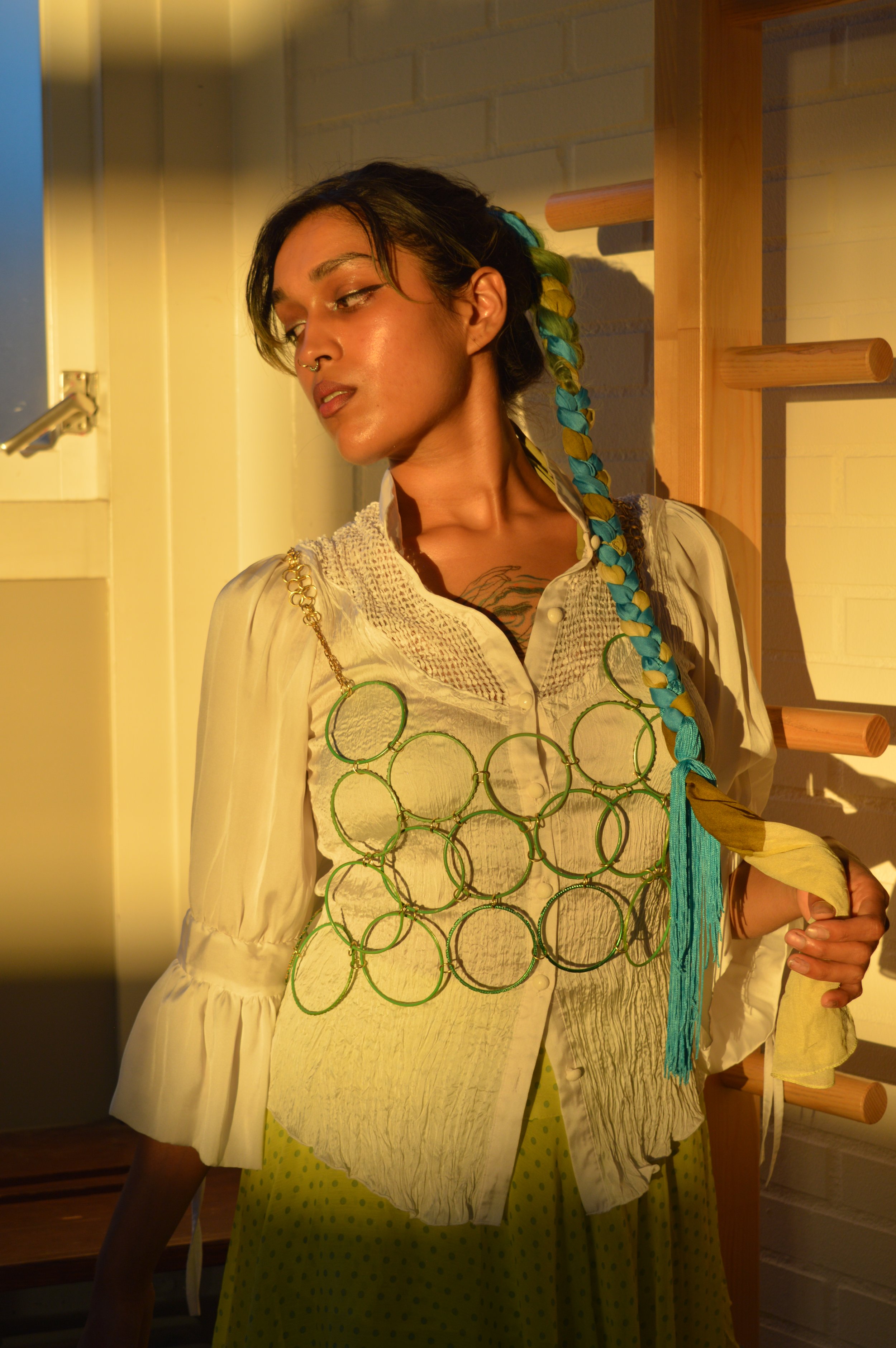 A person with medium skin tone, dark hair with colorful extensions, wearing a cream blouse, a green circular jewelry necklace, and a yellow polka dot skirt, stands against a white brick wall and wooden ladder, bathed in warm sunlight.