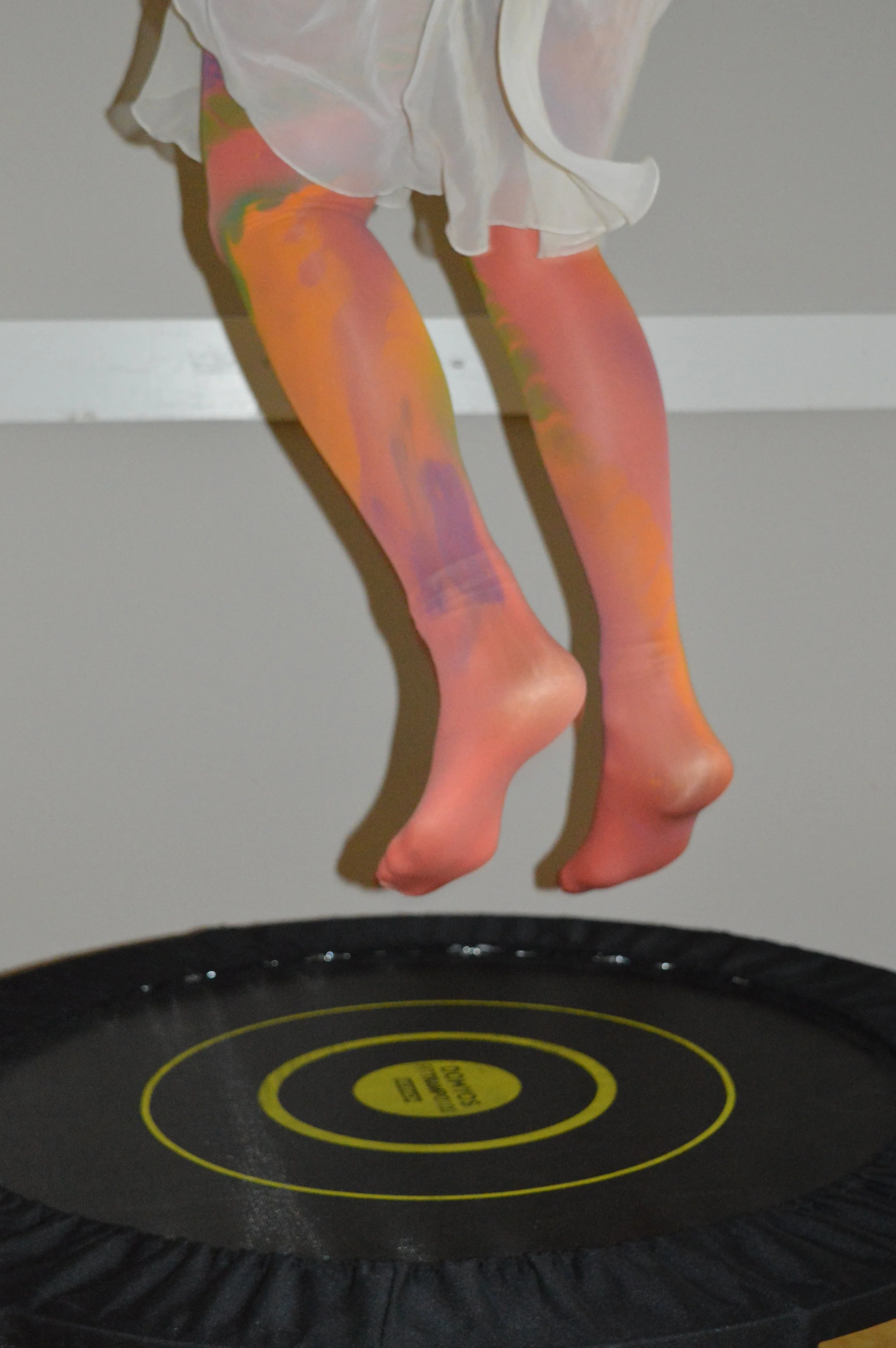 Close-up of pink and orange tights on a person's legs bouncing on a trampoline, with a white dress or skirt partially visible at the top of the image.