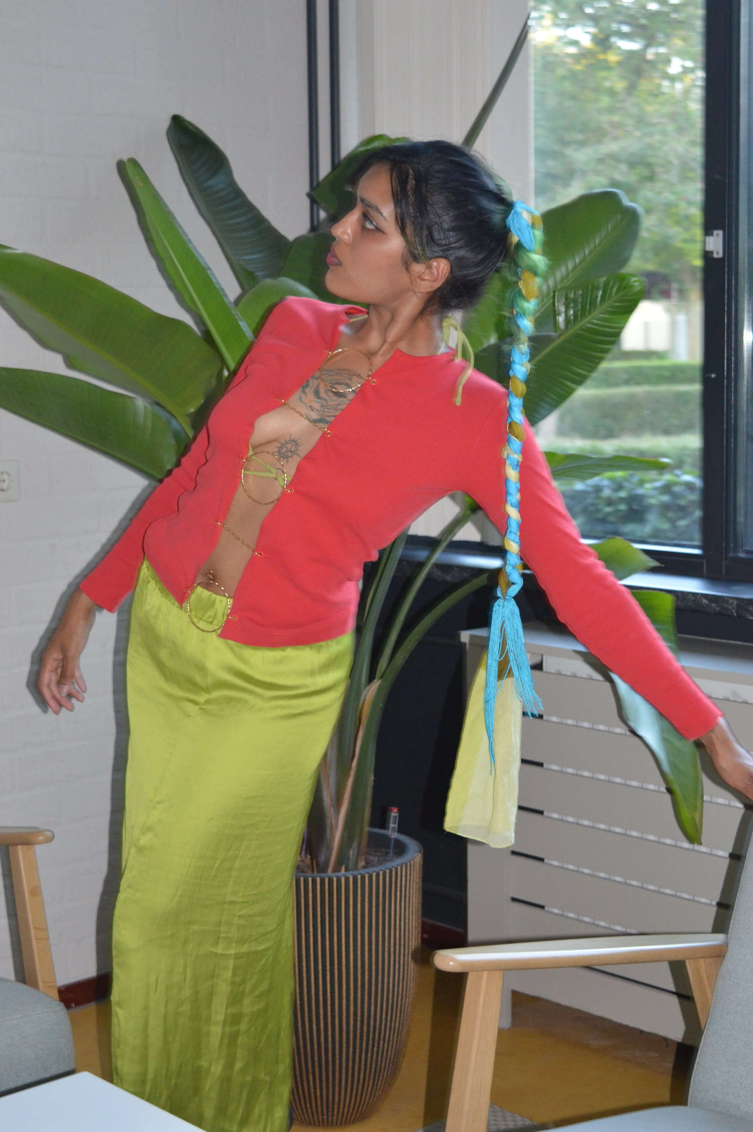 Woman with long braid wearing a red top and green skirt, standing near a large plant indoors.