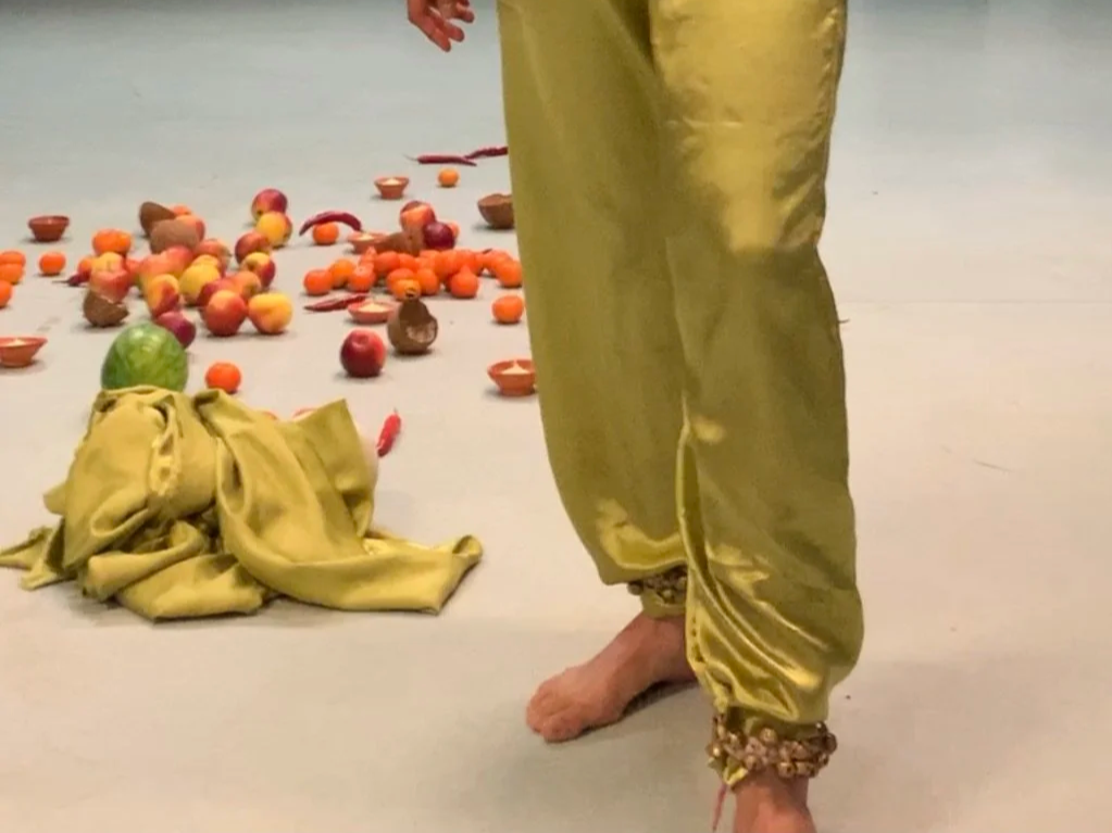 Person standing barefoot wearing shiny yellow pants with an Indian chain around the ankle, surrounded by scattered apples and other fruits, with some bowls / thali's and a green cloth on the floor.