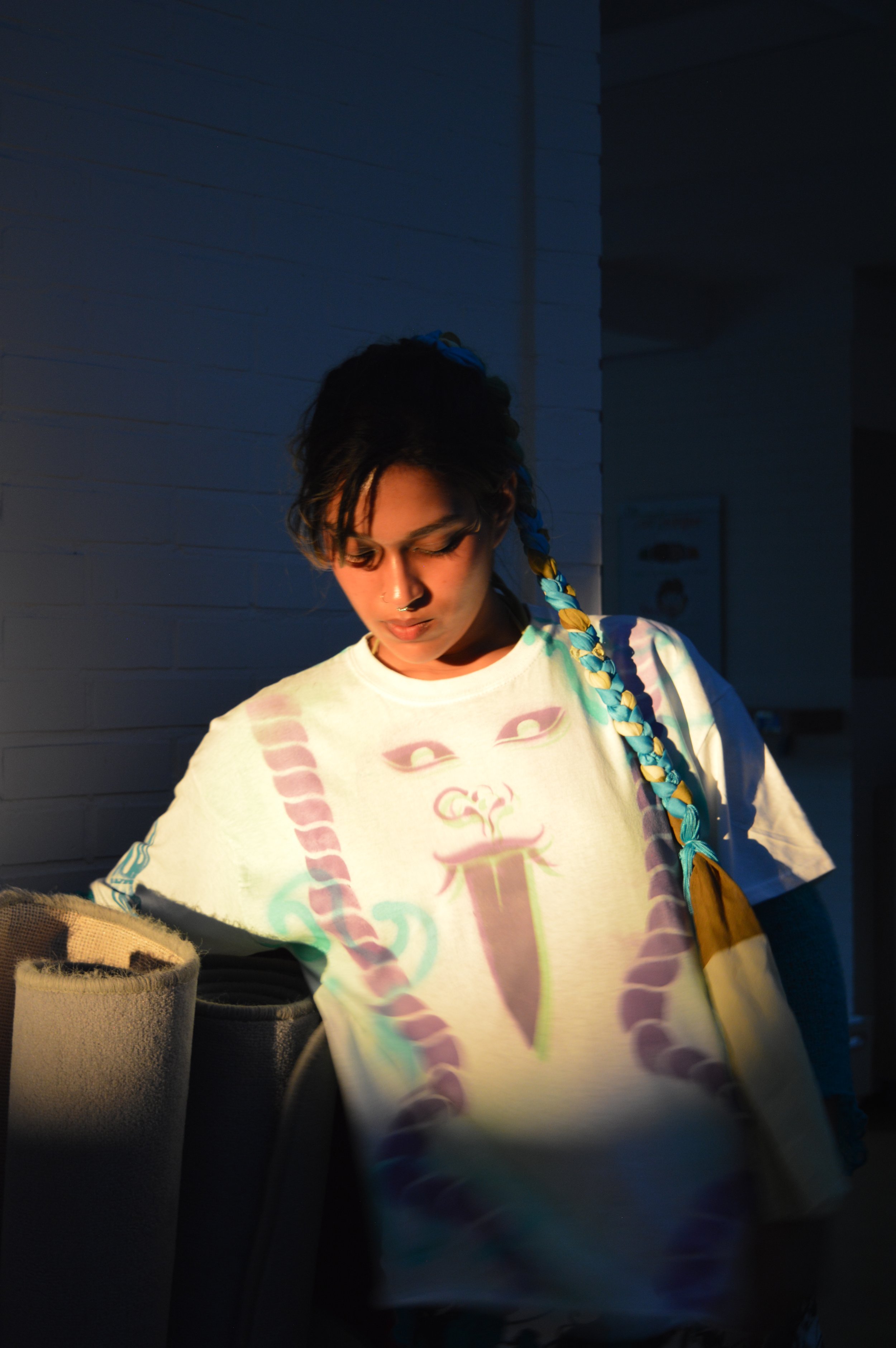 Young woman with braided hair and septum piercing looking down, illuminated by colorful projection light, standing next to tote bags against a brick wall.