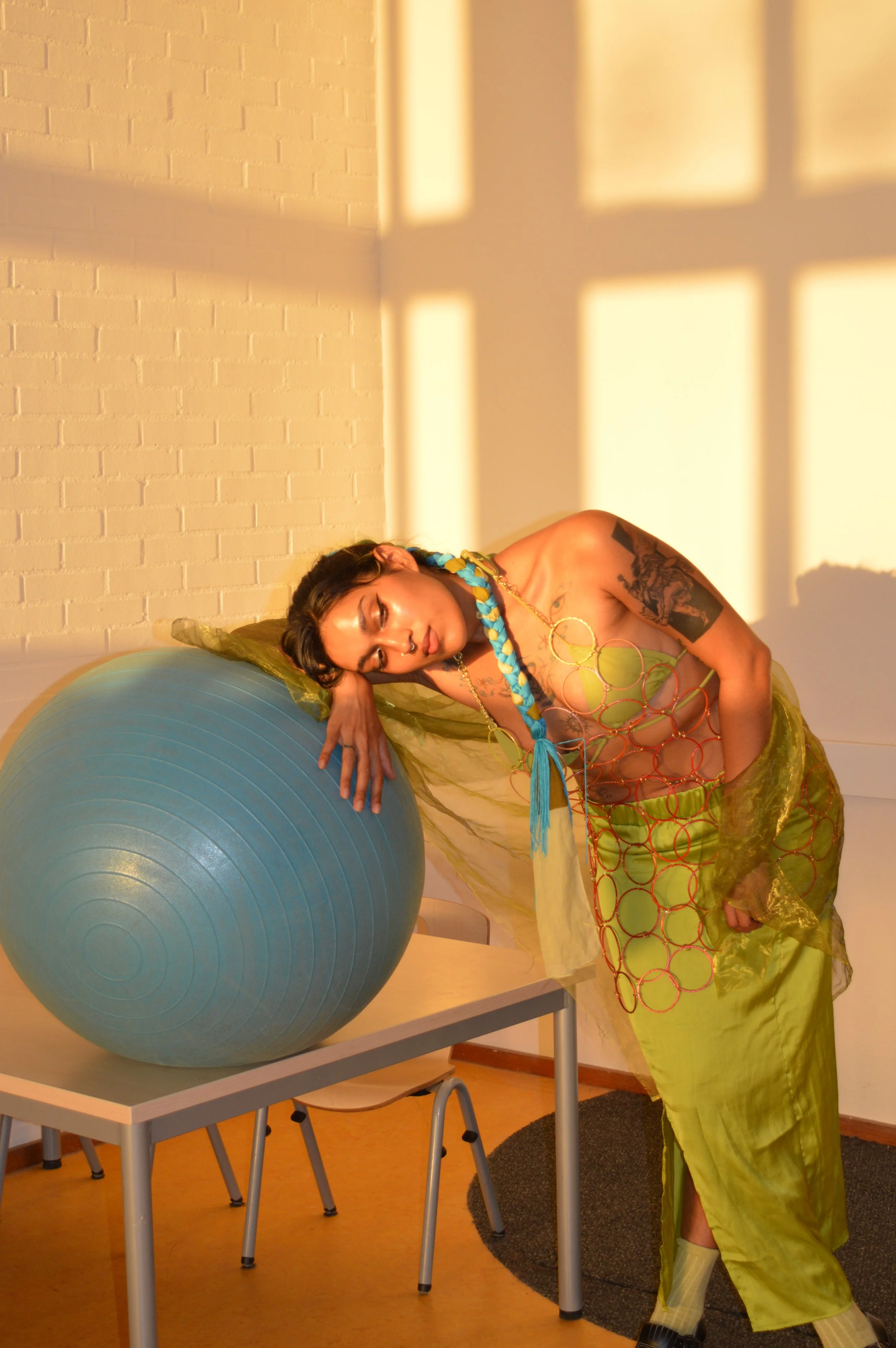 A woman with dark hair, tattoos, and colorful accessories resting her head and arm on a large blue exercise ball, in a room with sunlight and shadow patterns on the wall.
