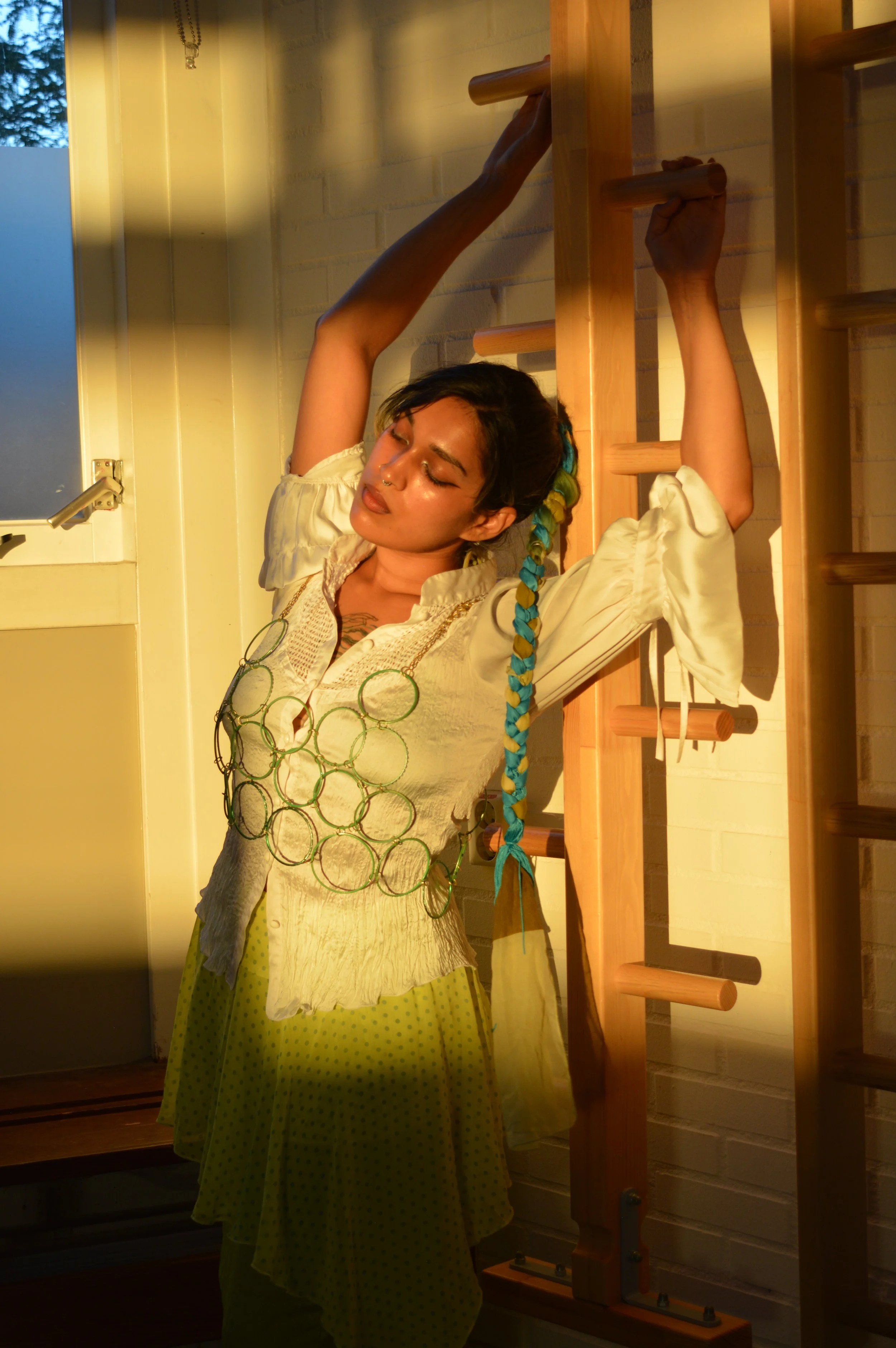 A woman with a rainbow braid and a green necklace is leaning against gym wall bars near a door, with sunlight illuminating her face and upper body.