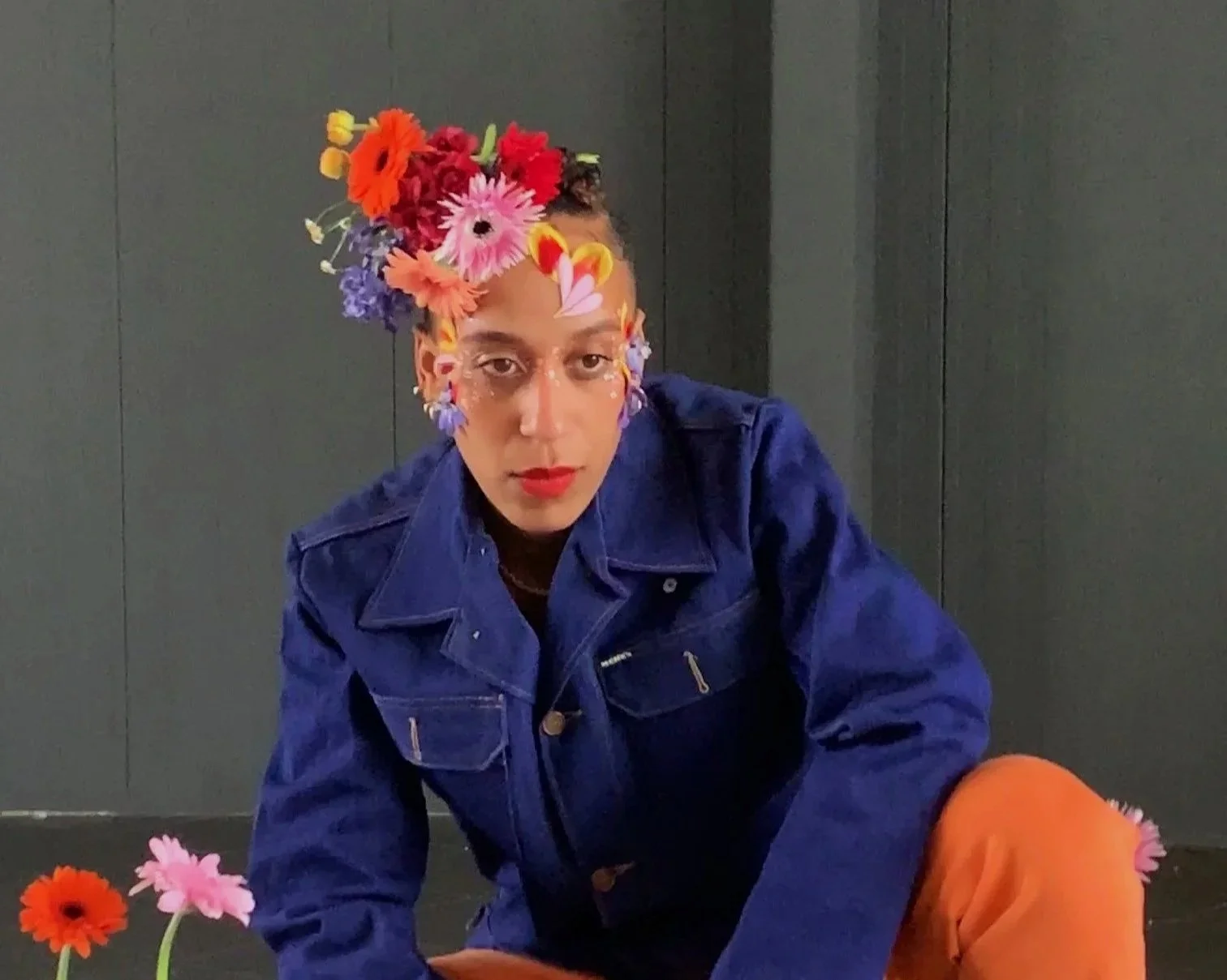 Person wearing a blue jacket and orange pants with colorful flowers on their head and face, sitting against a dark wall.