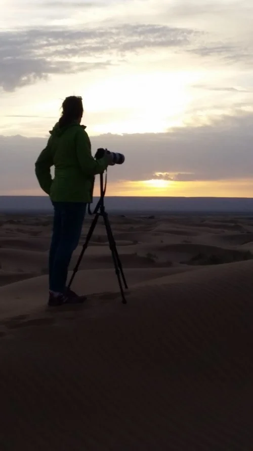 Silhouette of a person in a green jacket using a camera on a tripod at sunset in a desert landscape.