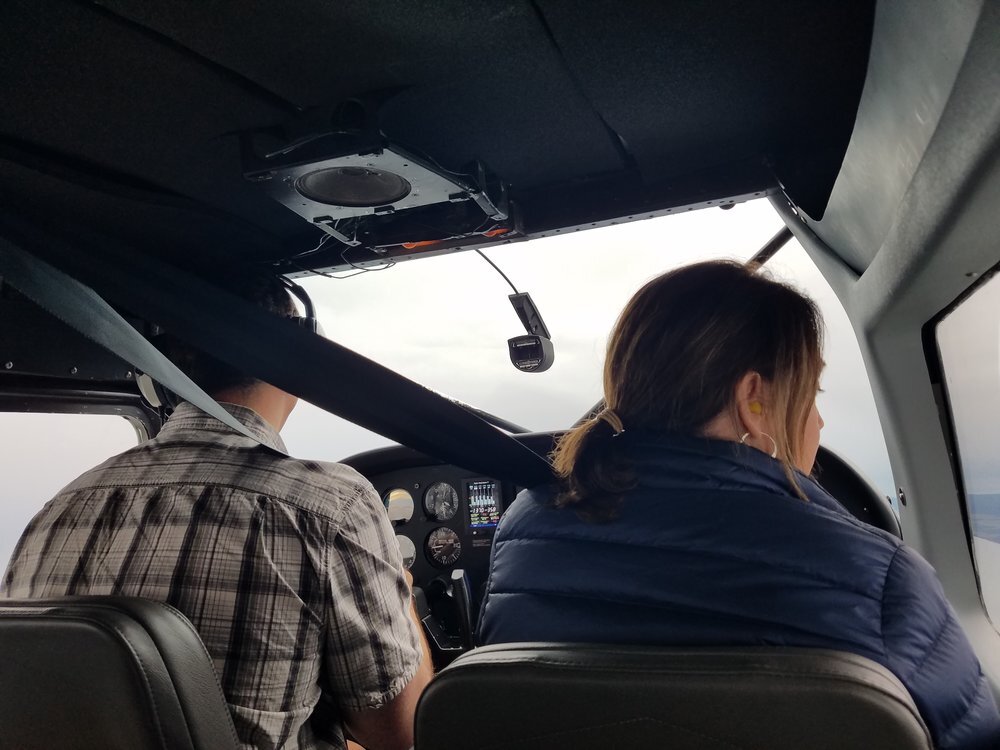 Interior view of a small aircraft cockpit with two people seated and wearing headsets. The cockpit displays various controls and instruments.