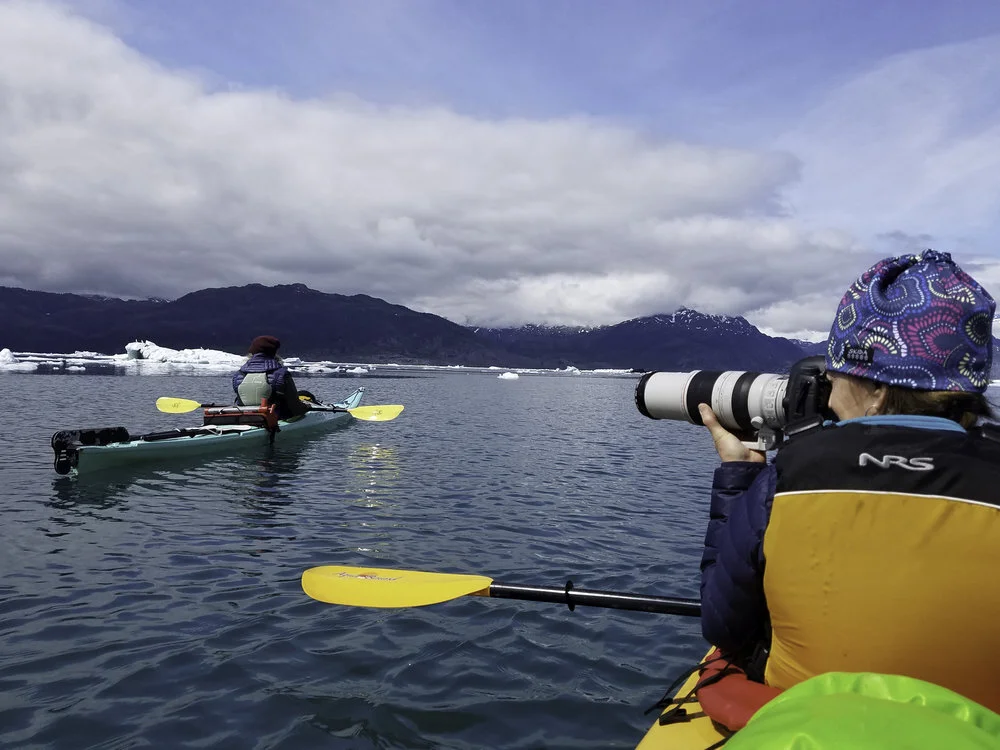 Two people kayaking in icy waters, one taking photographs with a large camera, surrounded by icebergs and snowy mountains under a cloudy sky.