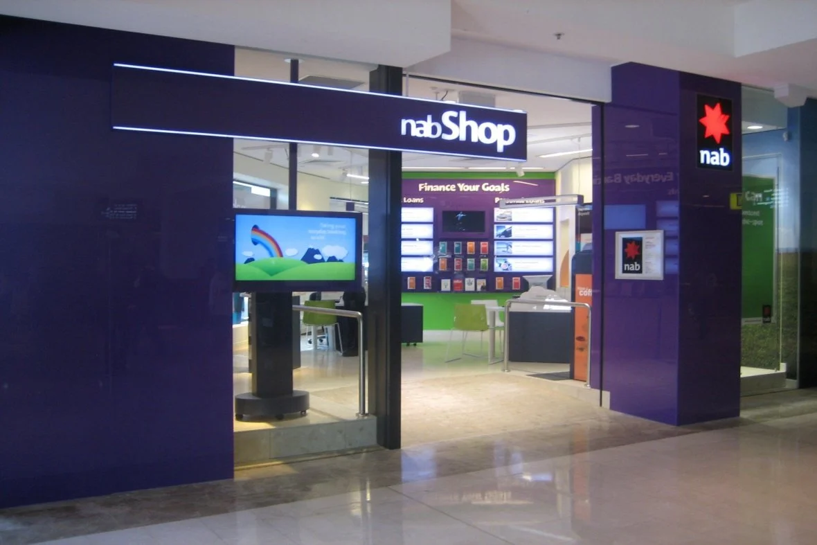 Entrance of a NAB Bank branch with purple exterior walls, a neon sign reading 'nabShop', a digital screen, and indoor seating area visible inside.