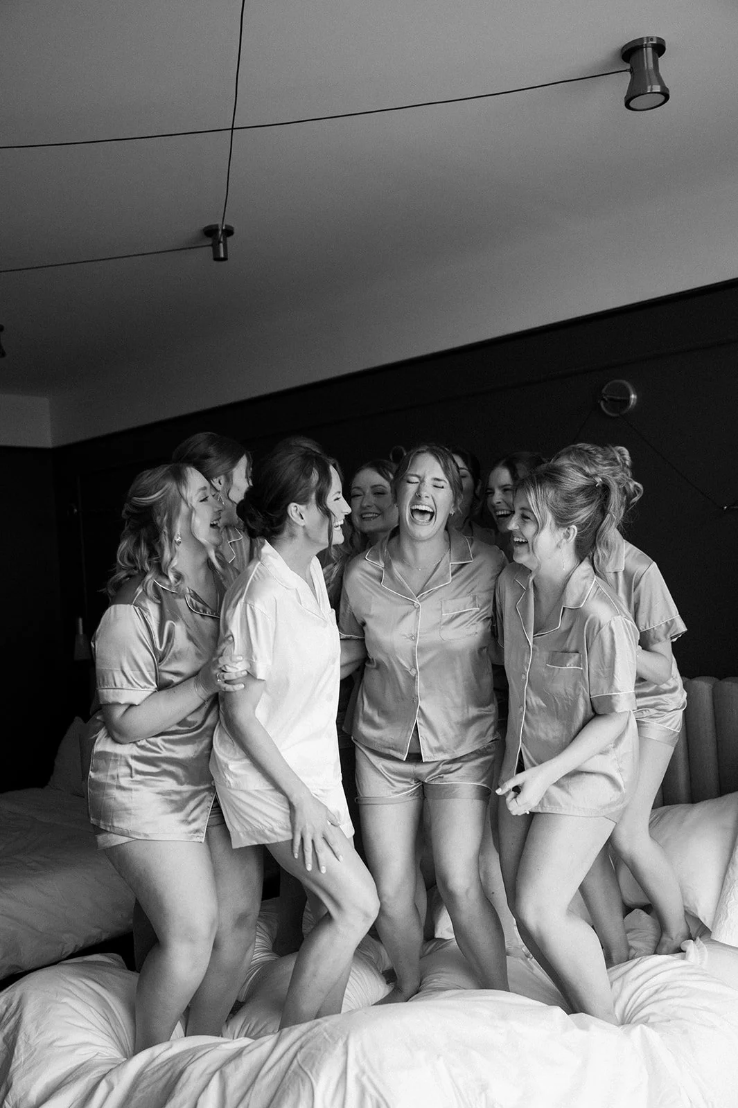 A group of women in pajamas laughing and dancing on a bed in a bedroom.