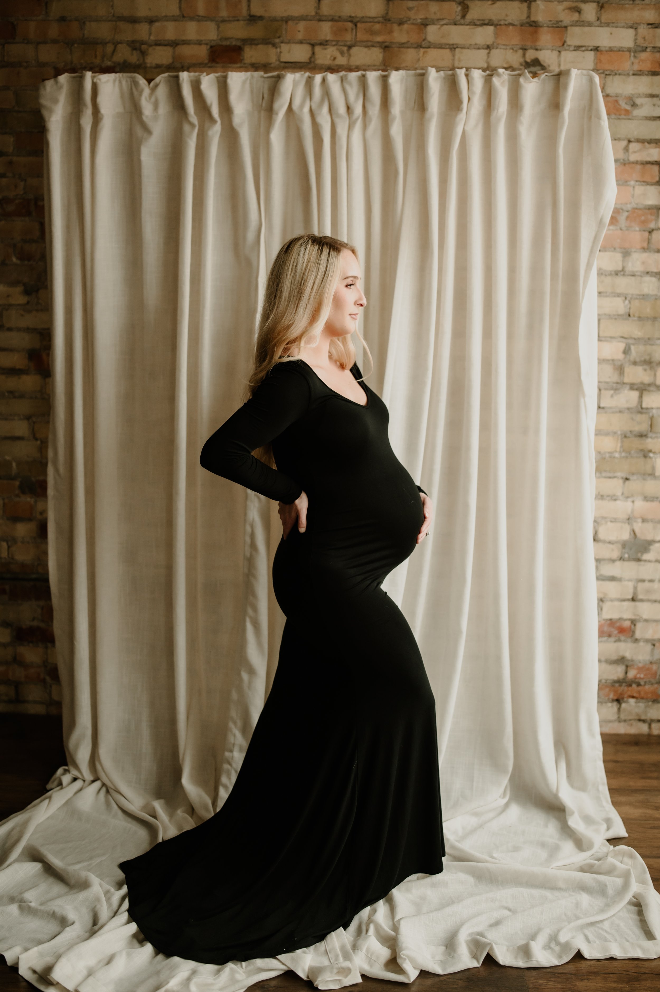 Pregnant woman in a black long dress standing sideways with hands on hips, posing in front of cream curtains and exposed brick wall.