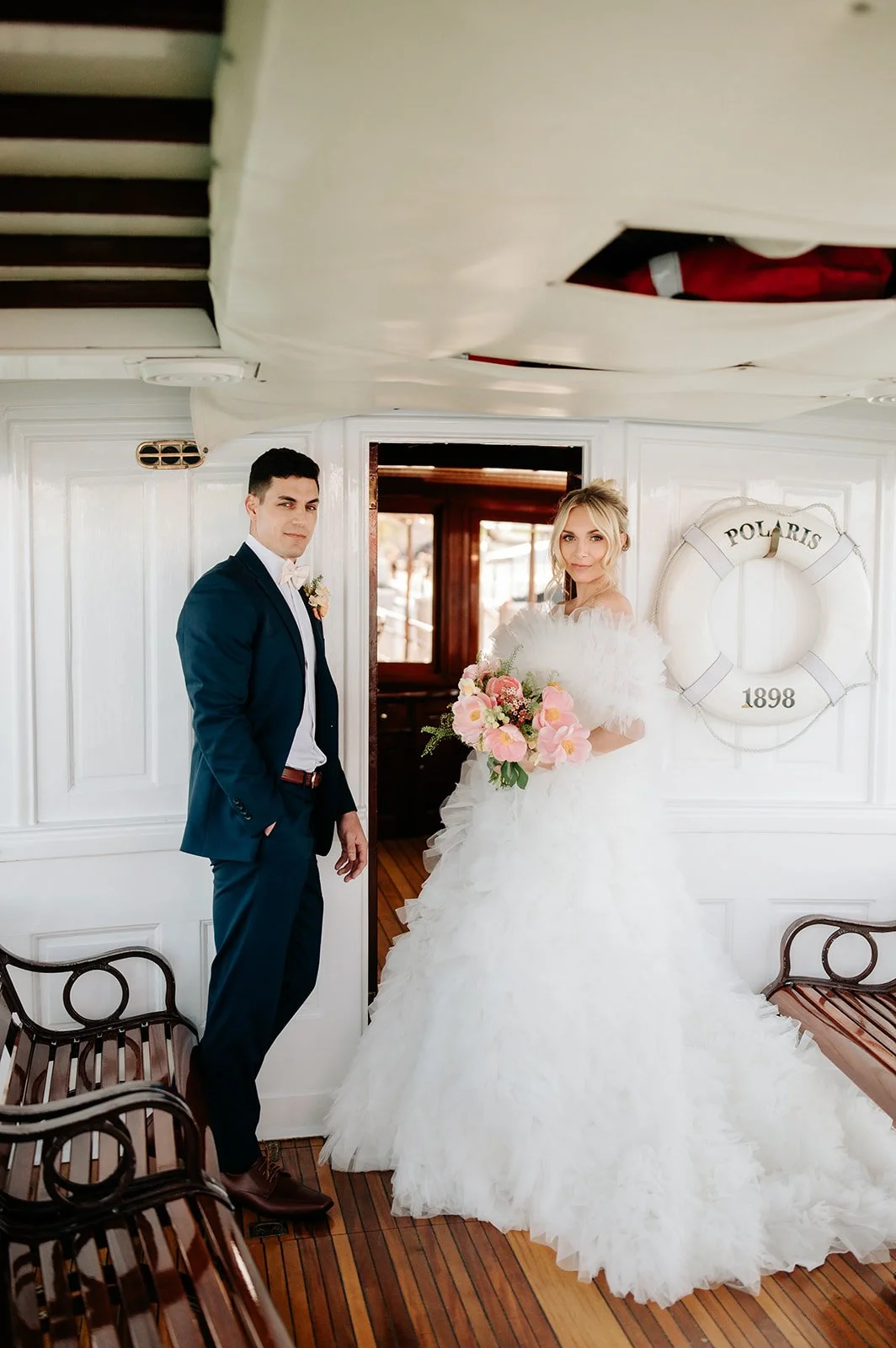 A bride and groom standing on a boat, dressed in wedding attire, with the bride holding a bouquet of pink and white flowers.