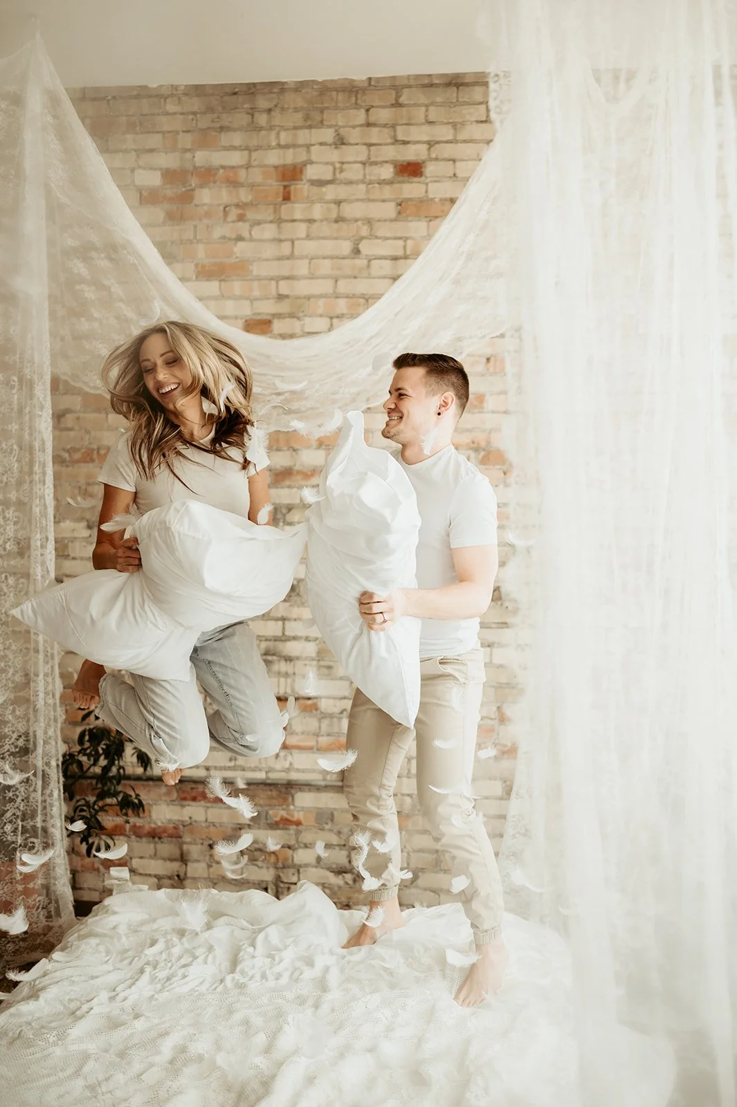 A smiling woman jumping on a bed holding a pillow, and a man standing on the bed holding a pillow, surrounded by falling feathers, in a bedroom with a brick wall and sheer curtains.