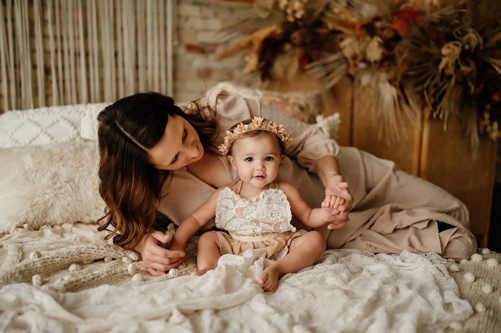 A woman and a young child lying on a bed, with the woman holding the child's hand. The girl is wearing a lace top and a floral headband, and the woman is dressed in a beige dress. The bed has cozy blankets and plush pillows, with a decorative wooden 