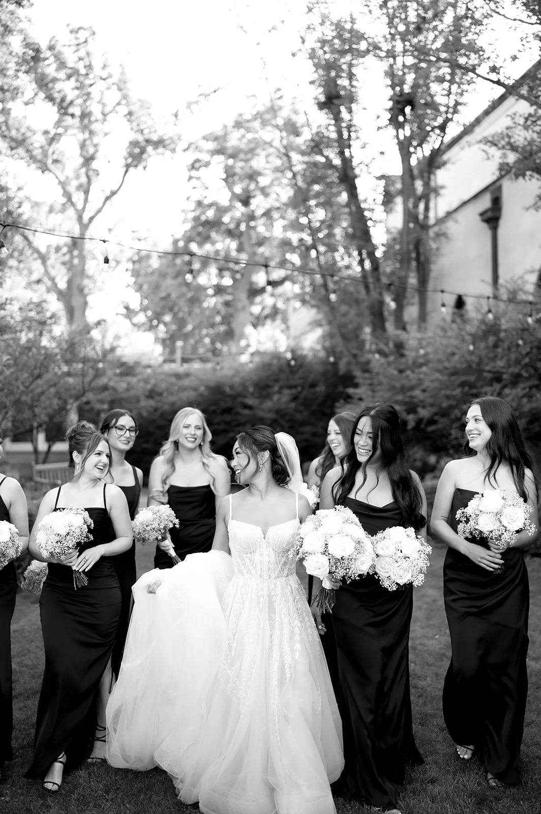 Black and white photo of a bride and her bridesmaids walking outdoors, smiling and carrying bouquets of flowers.