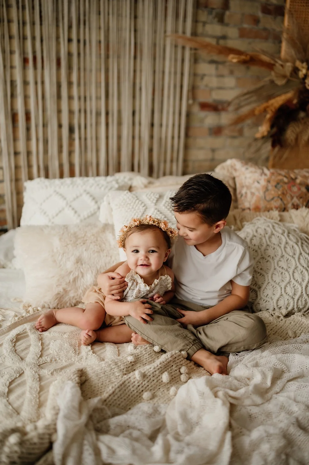 A young boy and a toddler girl sitting on a cozy bed with cream-colored blankets and pillows, in a warmly decorated room with a brick wall and dried floral arrangement in the background.