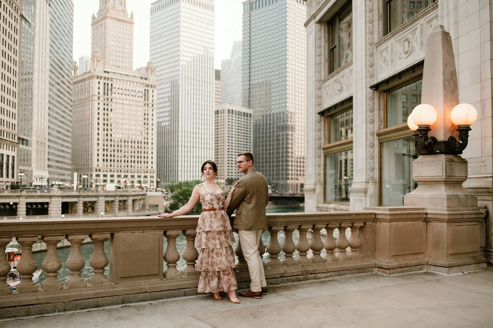 A young woman and man stand on a balcony overlooking a city with tall buildings. The woman is wearing a tiered beige floral dress, and the man is dressed in a brown blazer and white pants. There is a decorative lamp with five white globes mounted on 
