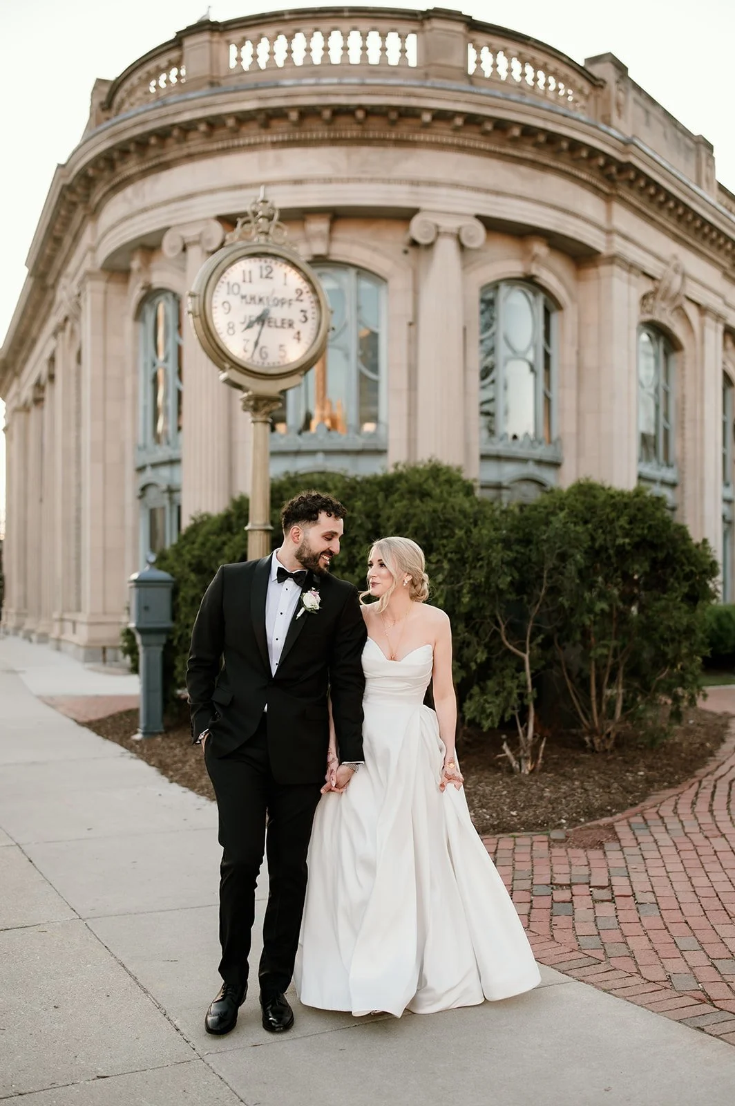A newlywed couple walking hand in hand outside with a historic building and a clock in the background, dressed in wedding attire, with the man in a tuxedo and the woman in a strapless white wedding gown.