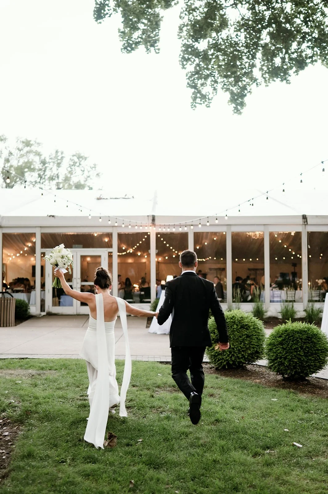 A bride in a white wedding dress and a groom in a black suit walking towards a decorated event tent, holding a bouquet and holding hands inside a wedding celebration outdoor venue.