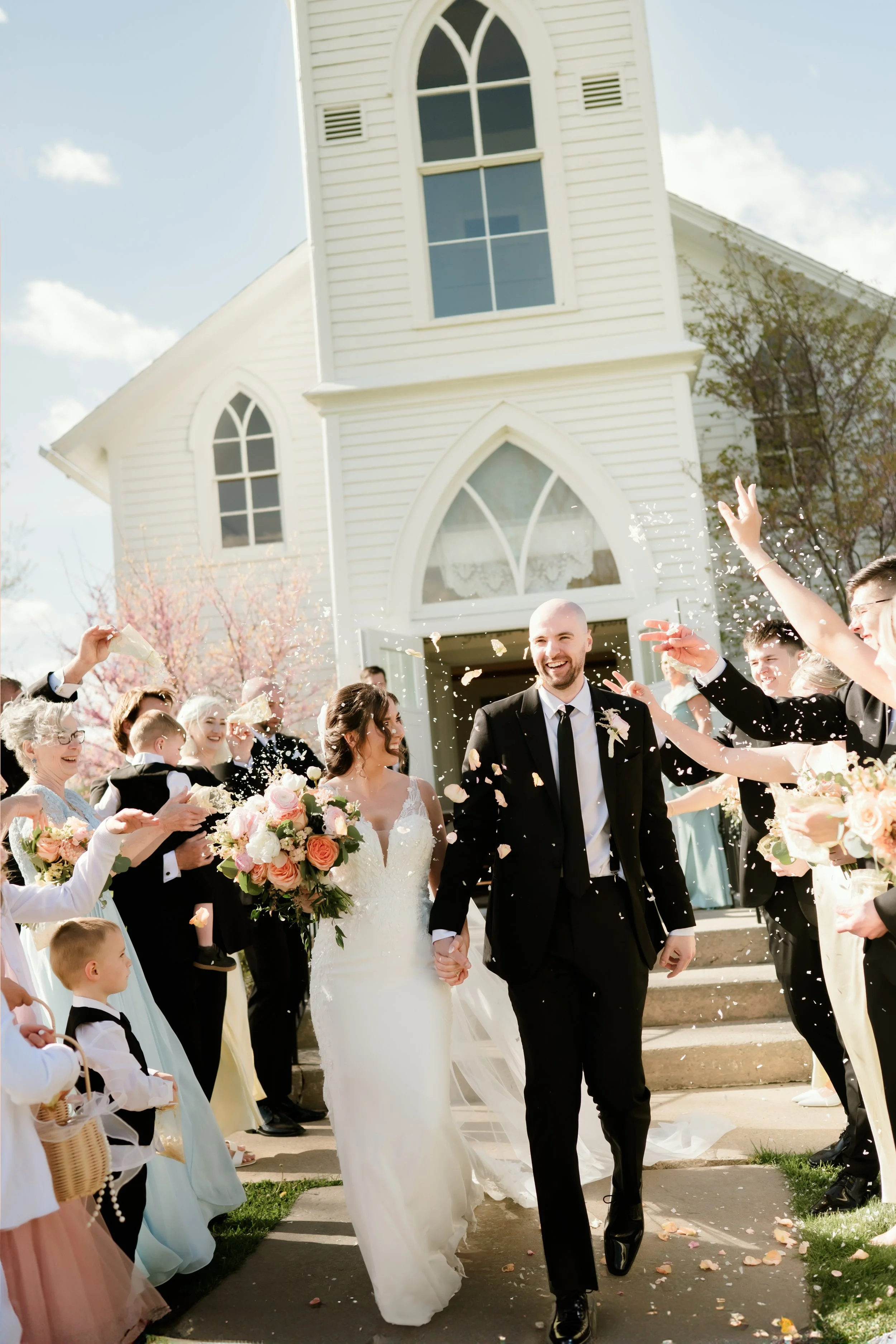 Wedding celebration outside a white church, with the bride and groom holding hands, surrounded by guests showering them with flower petals.
