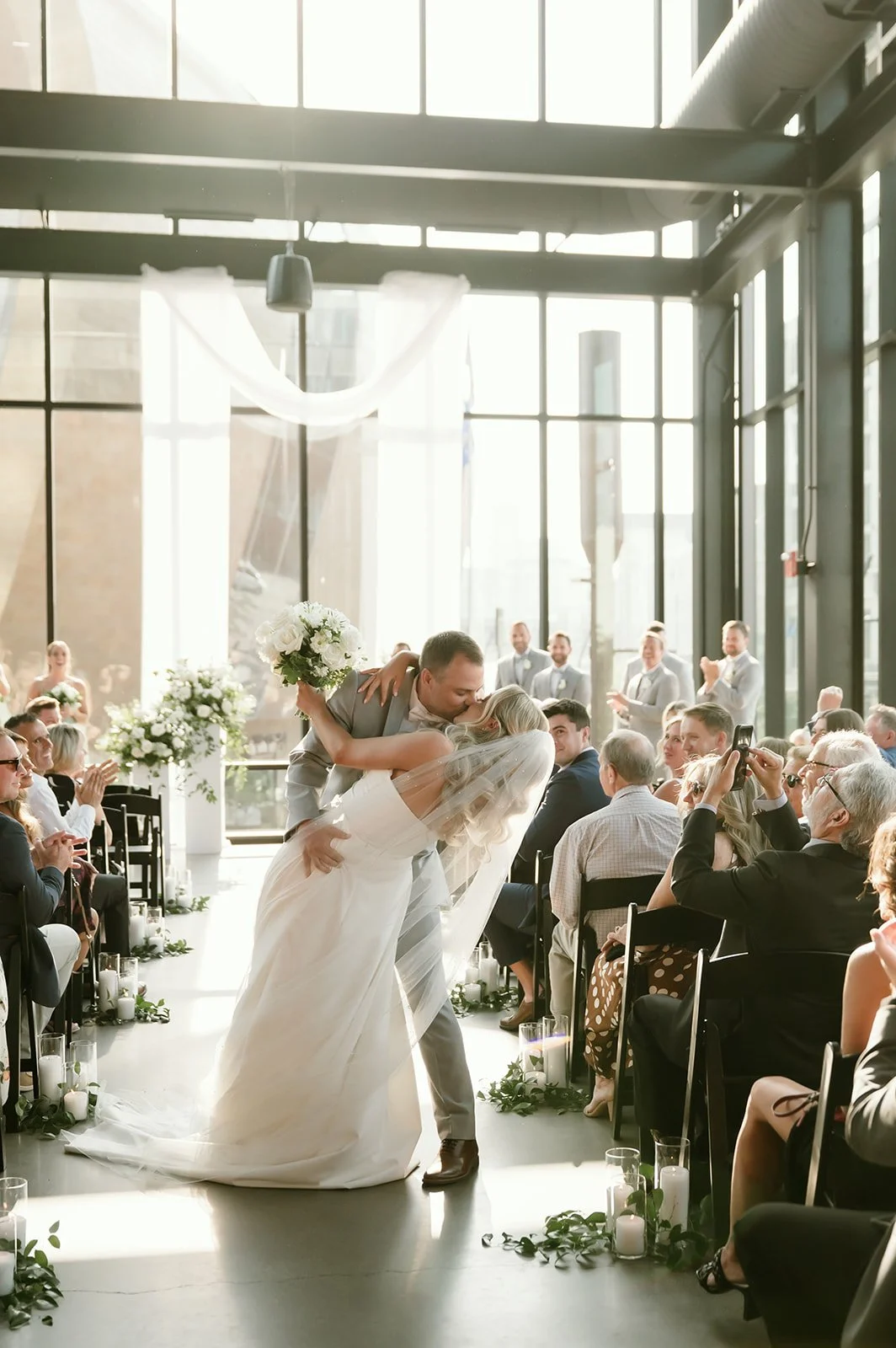 A bride and groom sharing a dance at their wedding ceremony inside a modern glass-walled venue, surrounded by seated guests and floral decorations.