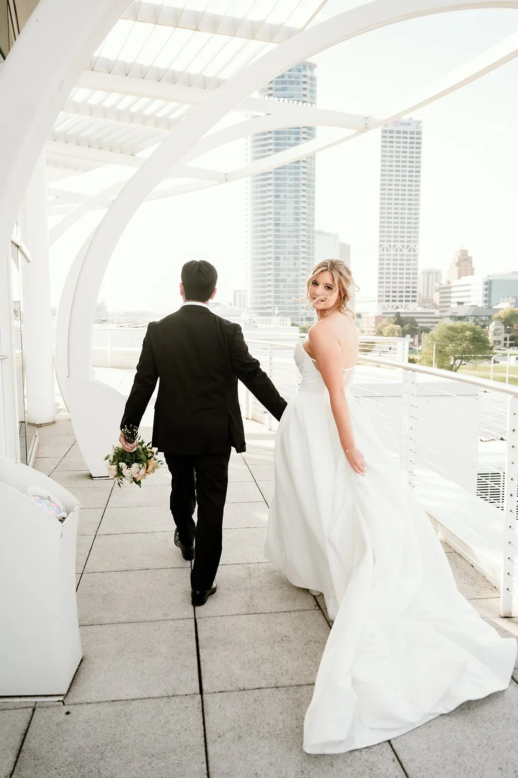 A newlywed couple holding hands on a rooftop balcony with a city skyline in the background. The bride is in a strapless white wedding gown, and the groom is in a black suit, holding a bouquet of flowers.