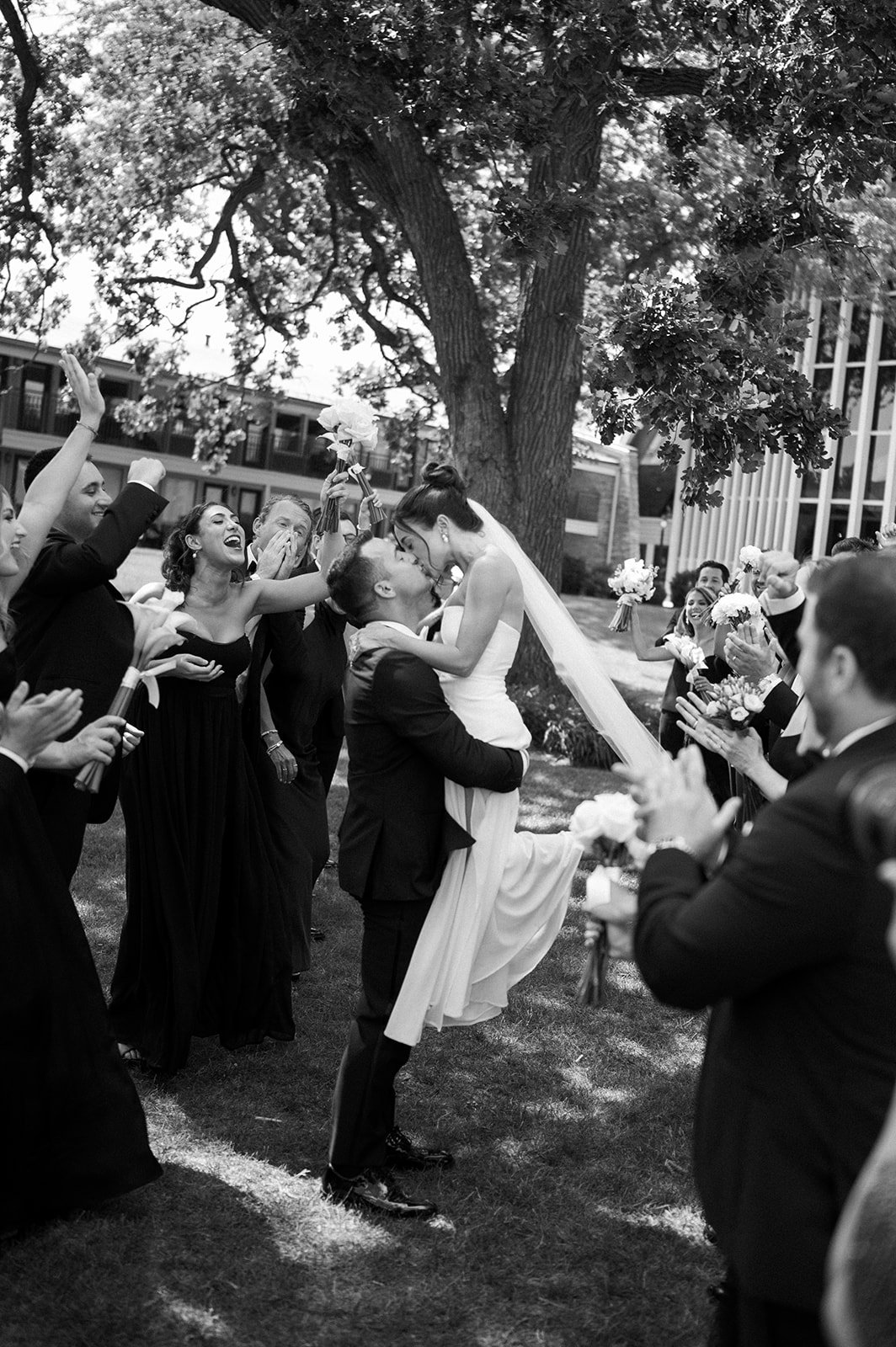 Black and white photograph of a wedding celebration showing a couple kissing surrounded by friends and family. The groom lifts the bride, and guests hold flowers and clap cheerfully outdoors beneath a large tree.