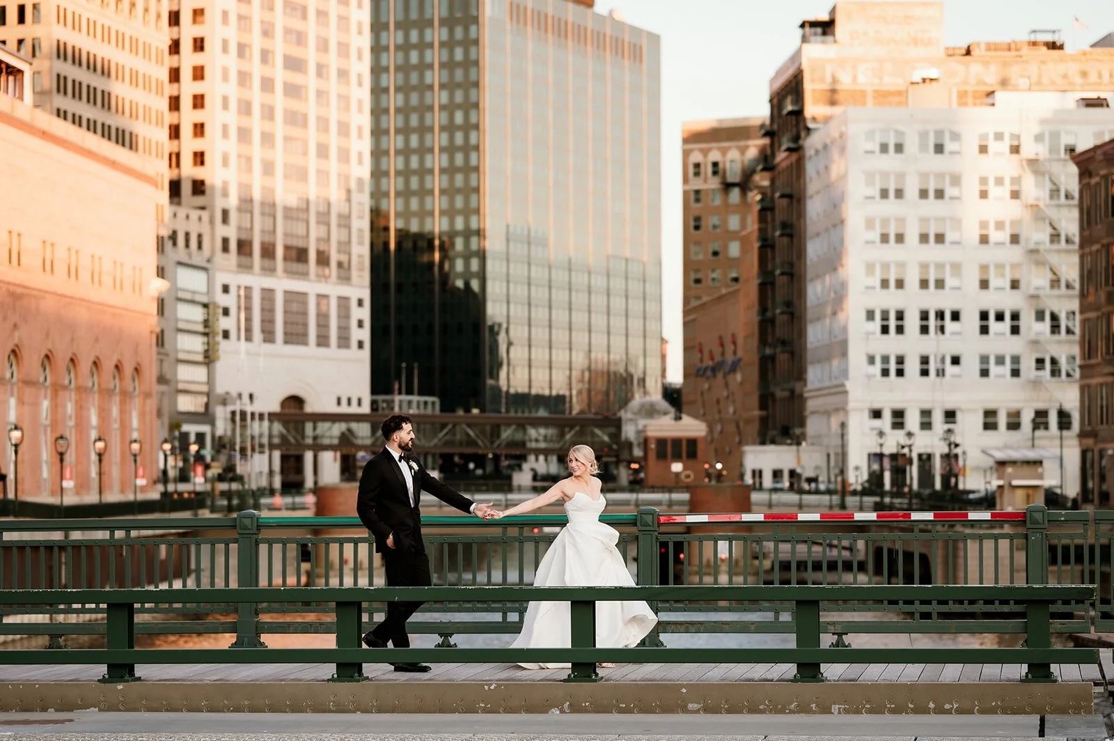A newlywed couple in formal attire holding hands on a city bridge with tall buildings in the background during sunset.