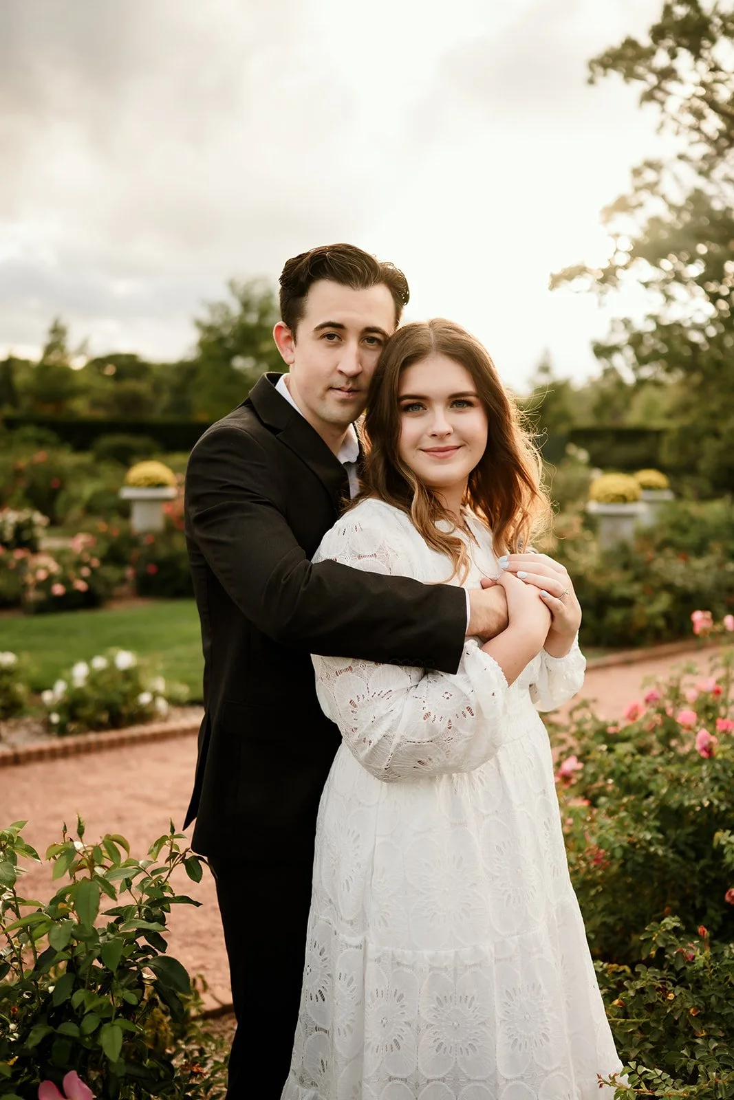 A young man and woman embrace in a garden with blooming roses, with a setting sun in the background.