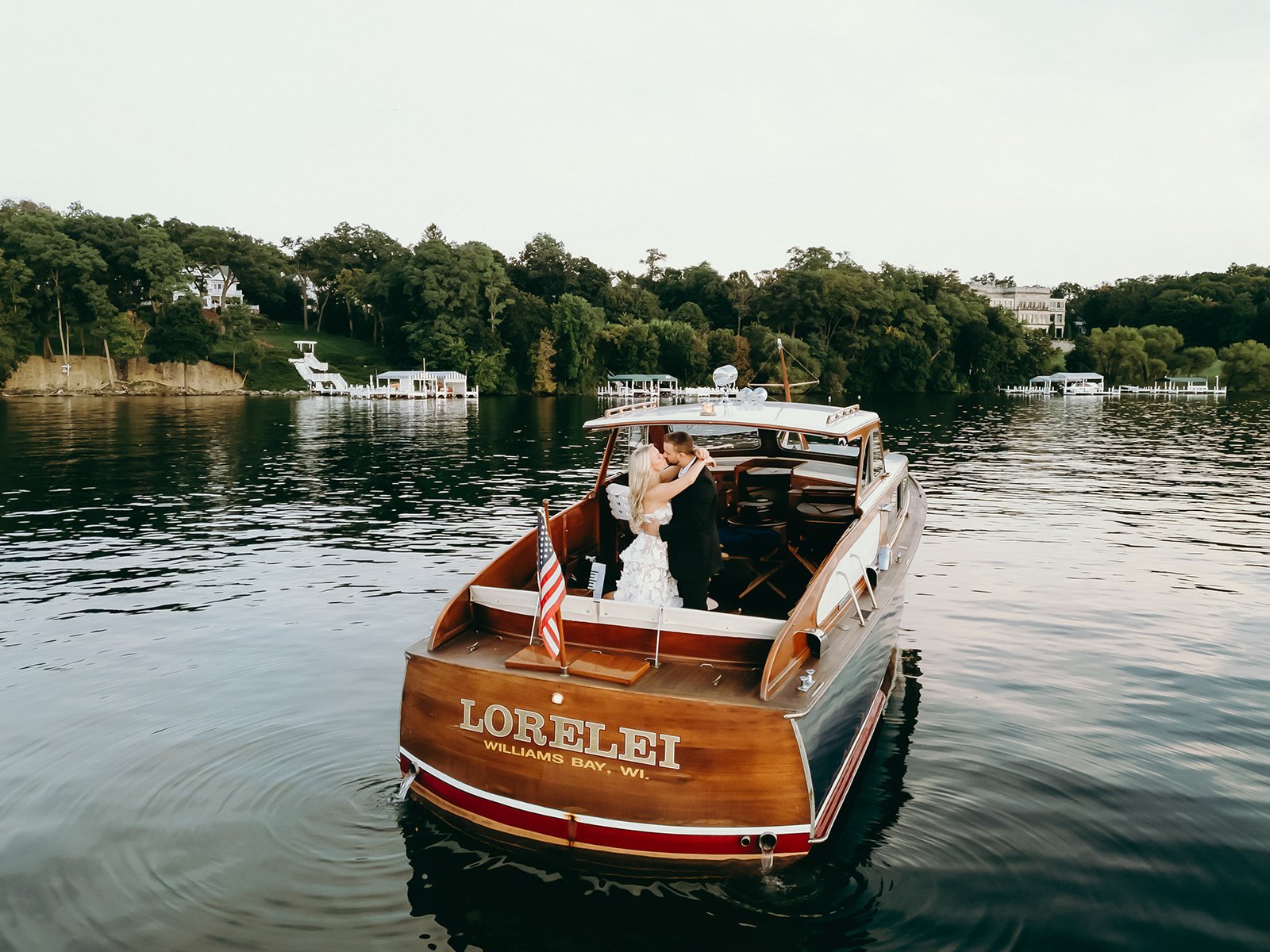 Two people sharing a kiss on a boat named Lorelei, with a scenic lake and forested shoreline in the background.
