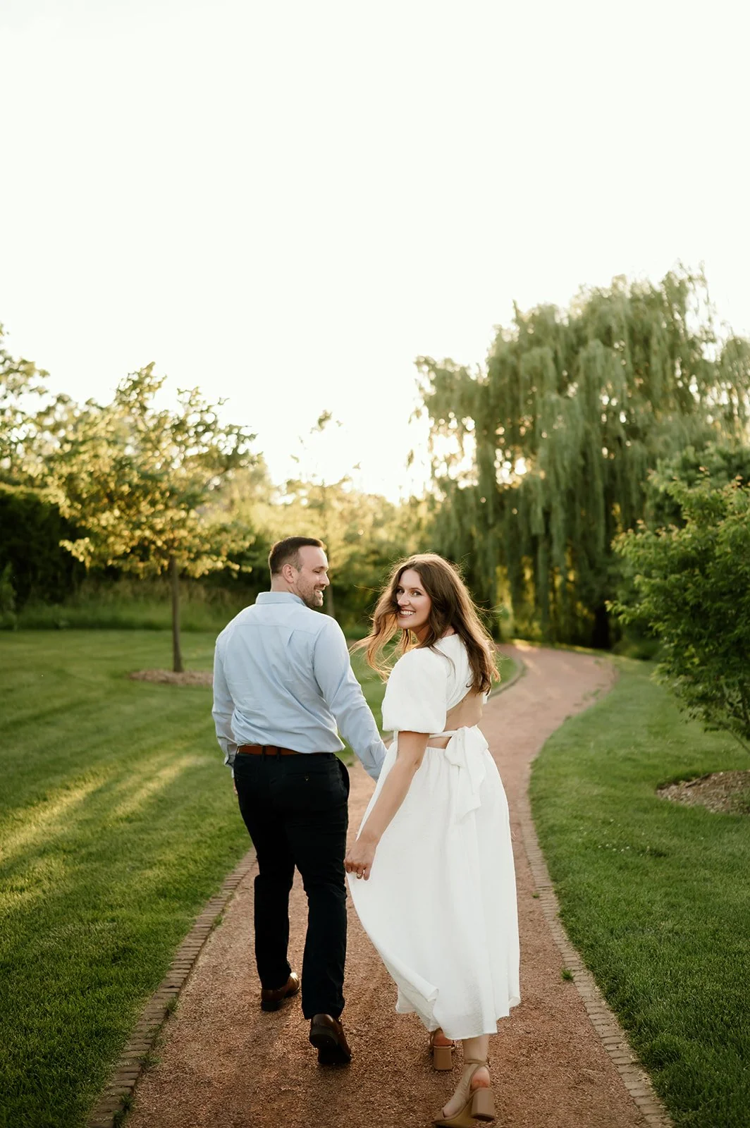 A couple walking hand in hand on a garden path, smiling and looking back at the camera during sunset.