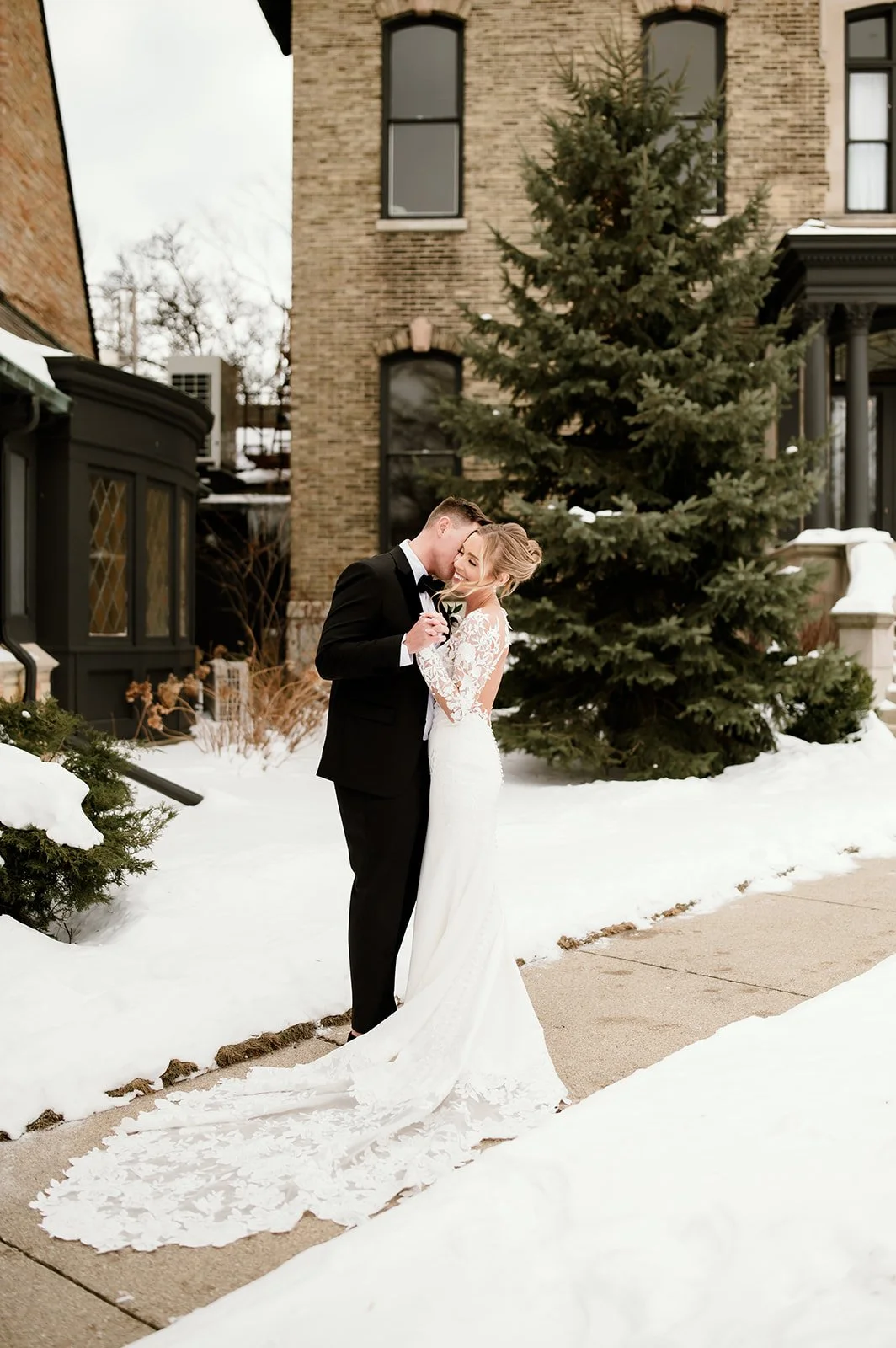 A bride and groom in wedding attire sharing a tender moment outdoors in the snow in front of a house with a large evergreen tree.