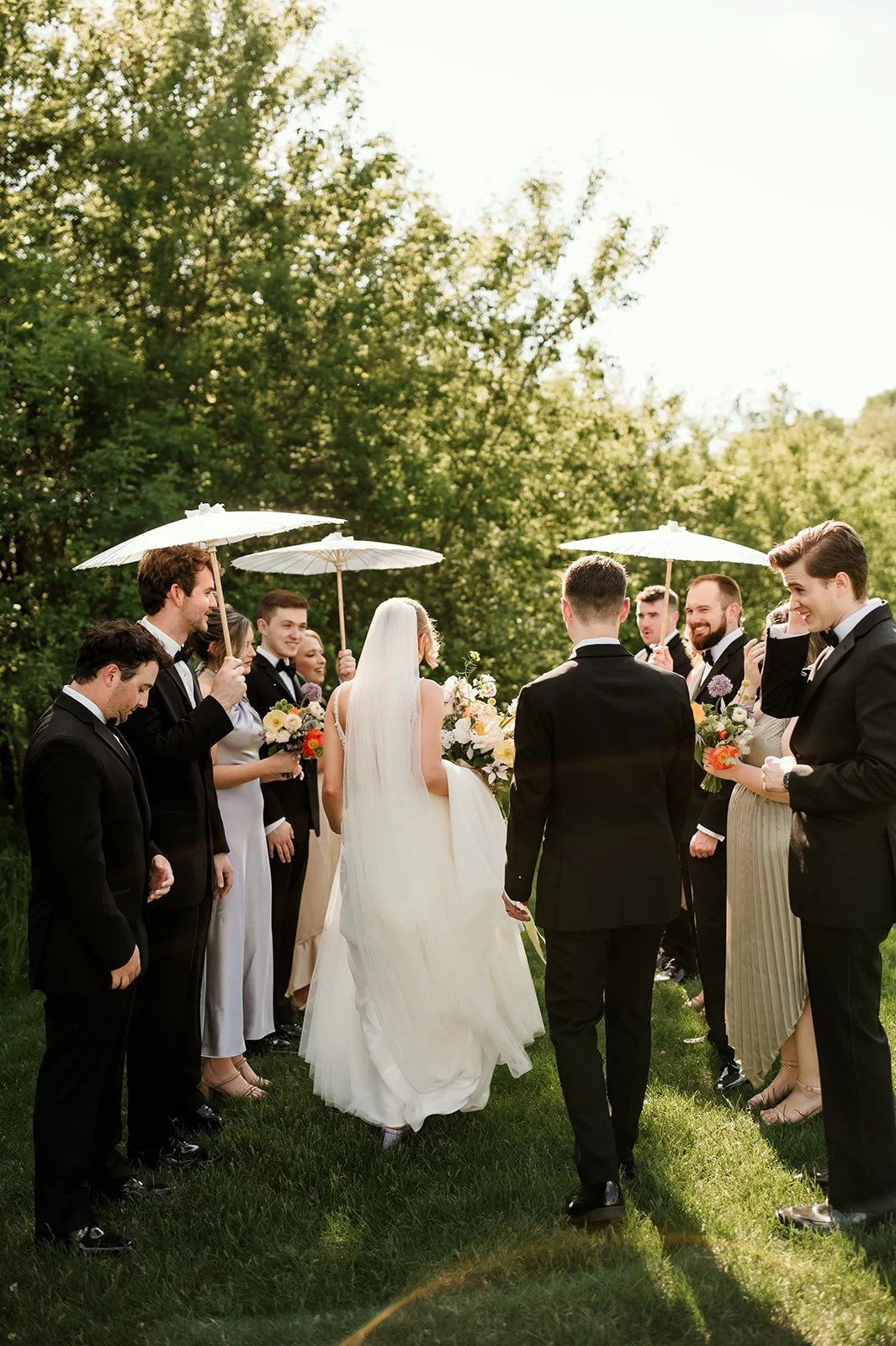 A bride and groom holding hands during their outdoor wedding ceremony, surrounded by their wedding party holding umbrellas, on a sunny day with green trees in the background.