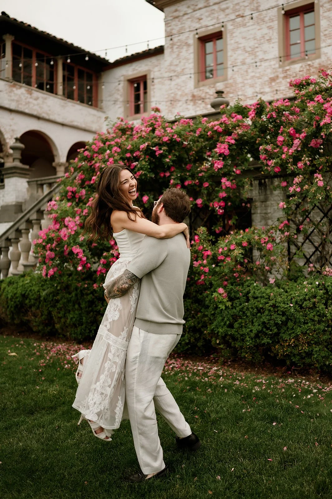 A man lifting a woman in a joyful outdoor setting with pink flowering bushes and a rustic building in the background.