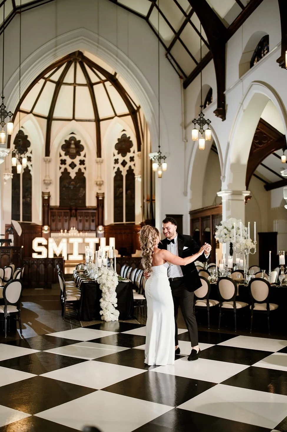 A bride and groom dancing in a decorated church wedding hall with high arched ceiling, chandeliers, and black and white checkered floor.