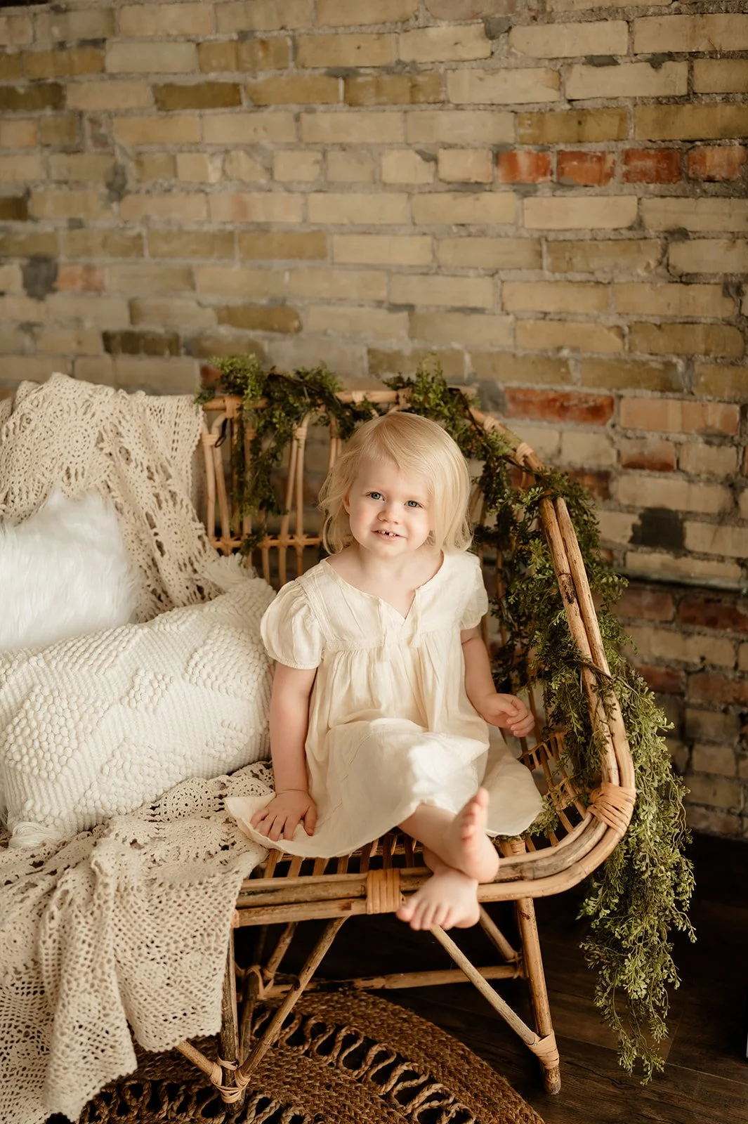 Young girl with blonde hair sitting on a rattan loveseat decorated with green foliage, against a brick wall, with cozy cream-colored pillows and crocheted blankets.