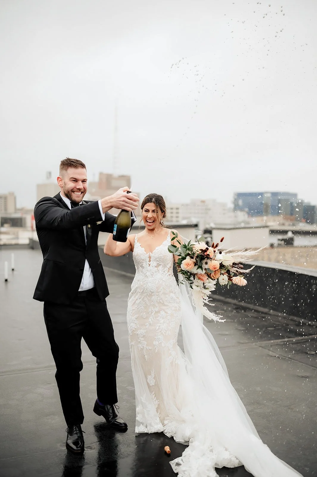 A wedding couple on a rooftop celebrating with champagne; the bride holds a bouquet; rain is falling.