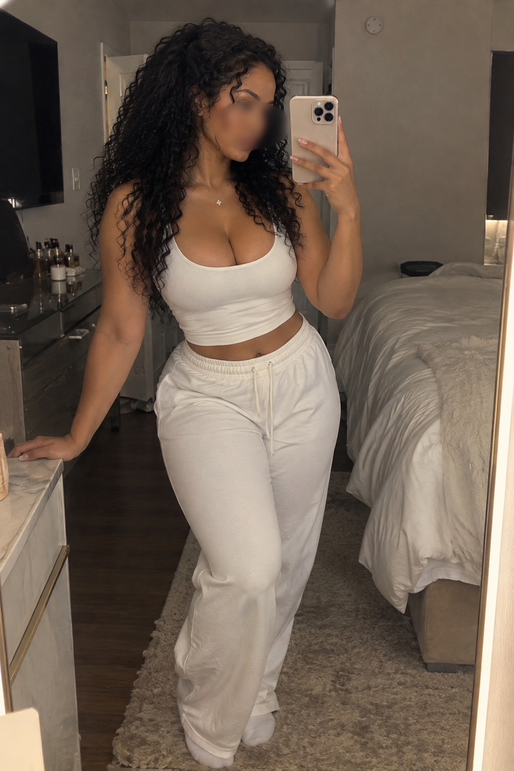 Woman with curly hair taking a mirror selfie in a bedroom, wearing a white crop top and white sweatpants.