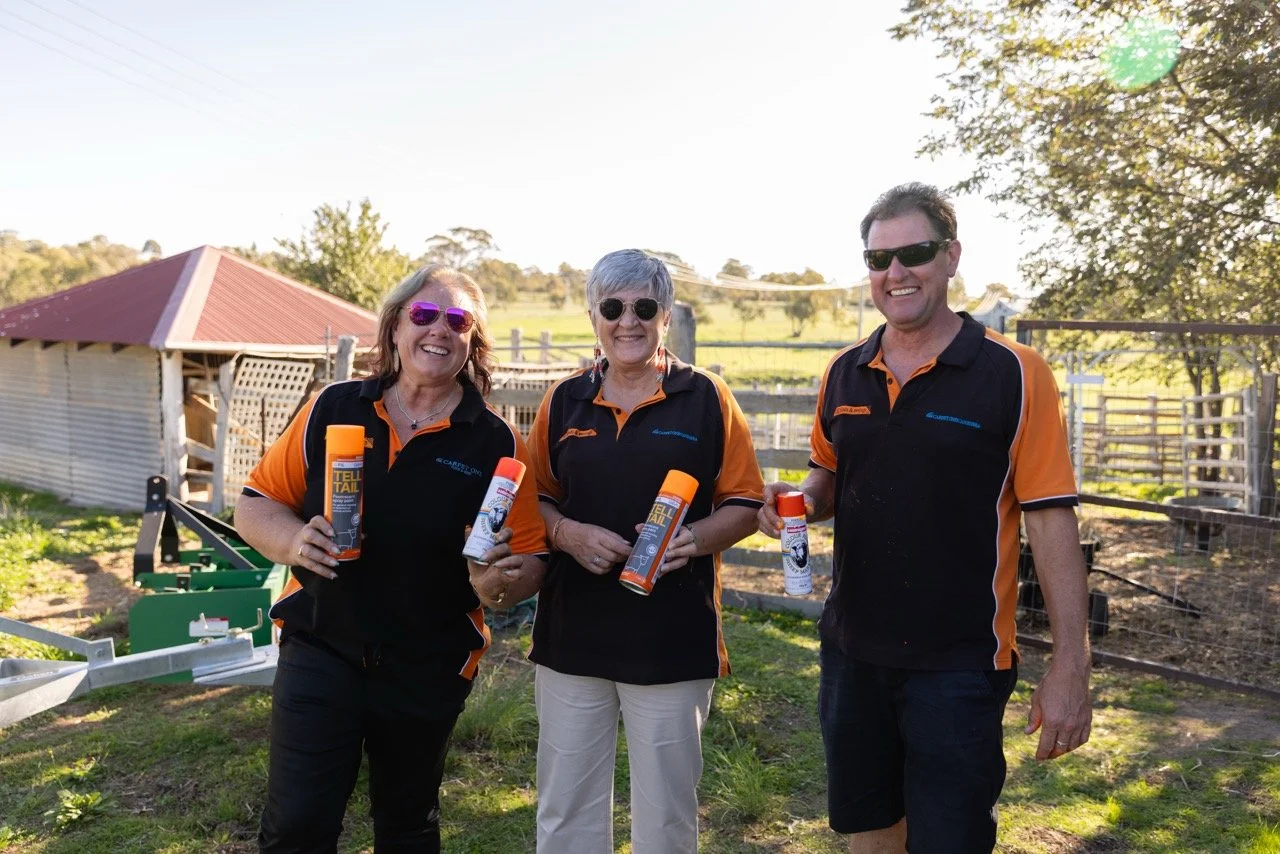 Three people from the CBI Blinds and Awnings team standing outdoors on a farm holding cans of spray, smiling, wearing matching black and orange uniforms, with a fence and trees in the background.