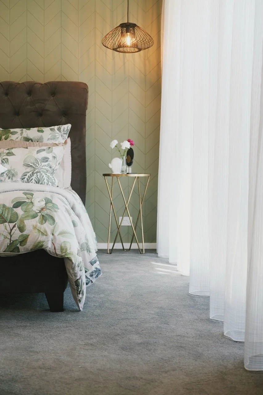 A bedroom corner with a bed, floral bedding, a tufted headboard, a side table with a vase of flowers, a geometric wire wall lamp, white sheer curtains, and a gray carpet.