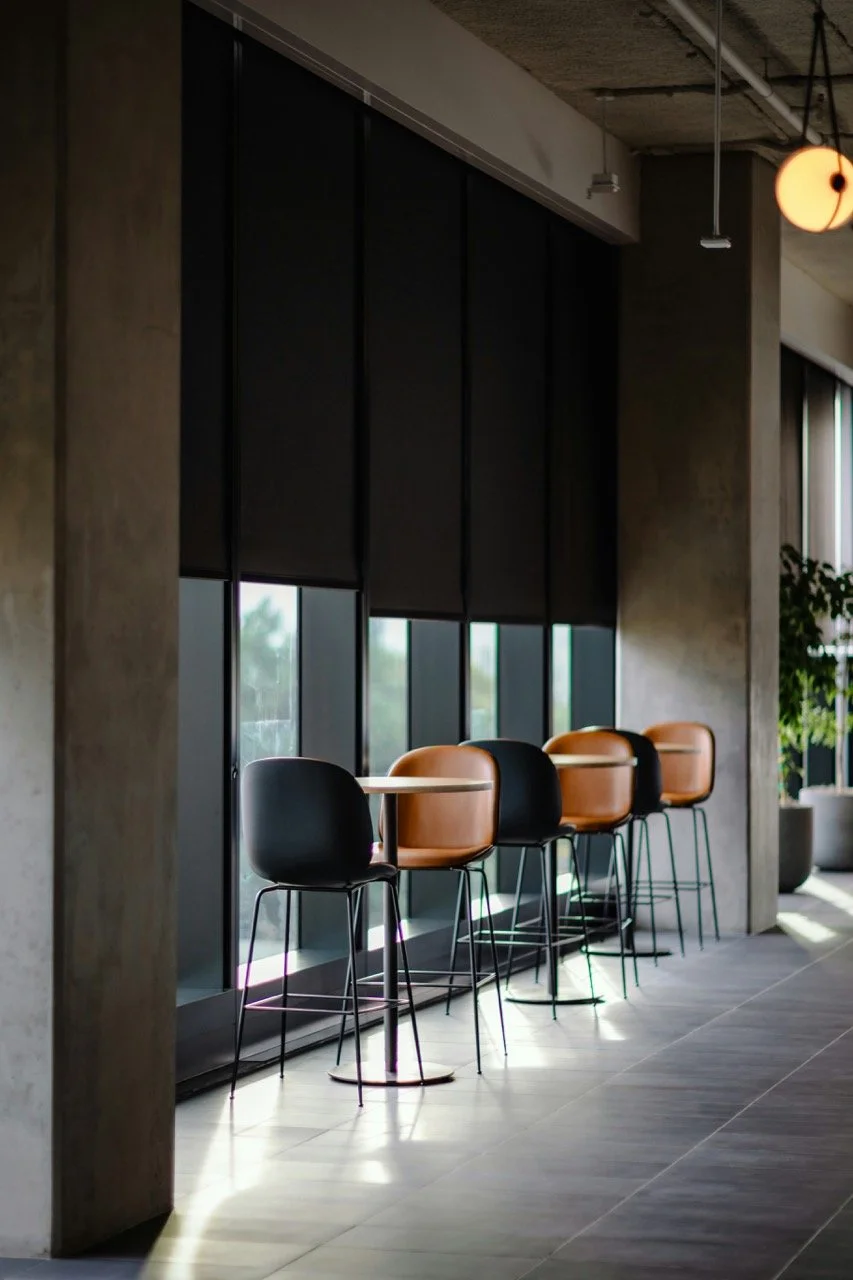 Interior of a modern cafe or lounge with a row of four bar stools, two black and two tan, placed along a windowed wall. The space features concrete walls, large windows with black roller blinds.