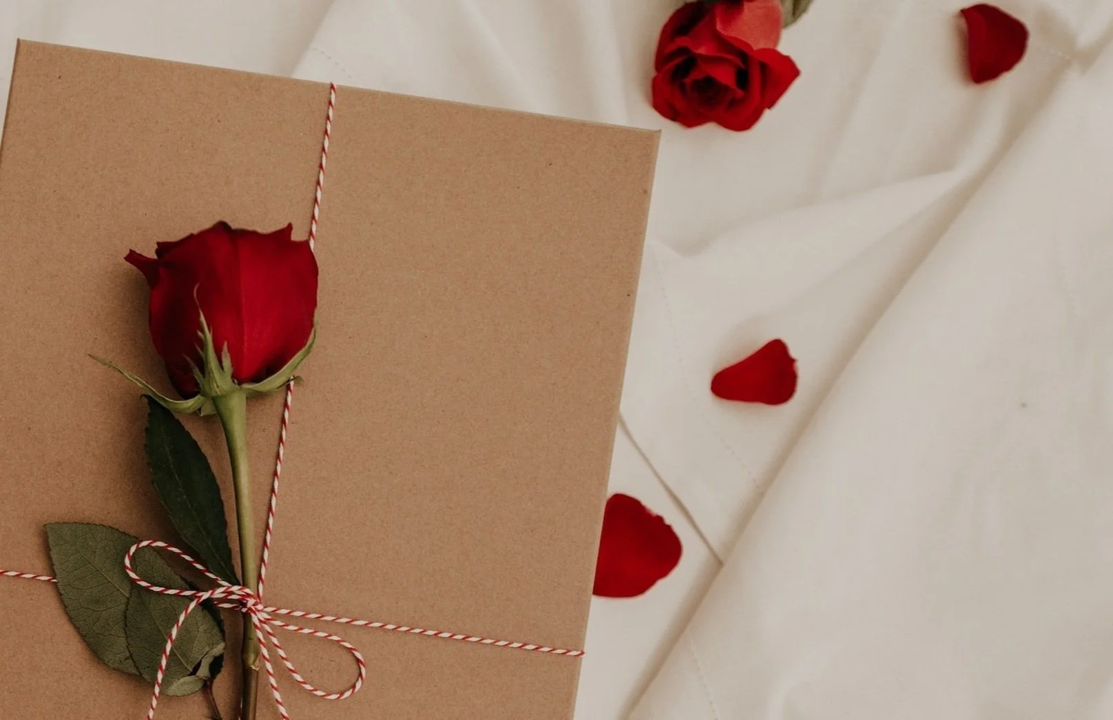 A brown gift box with a red rose and green leaves attached with red and white twine, lying on white fabric with scattered red rose petals.